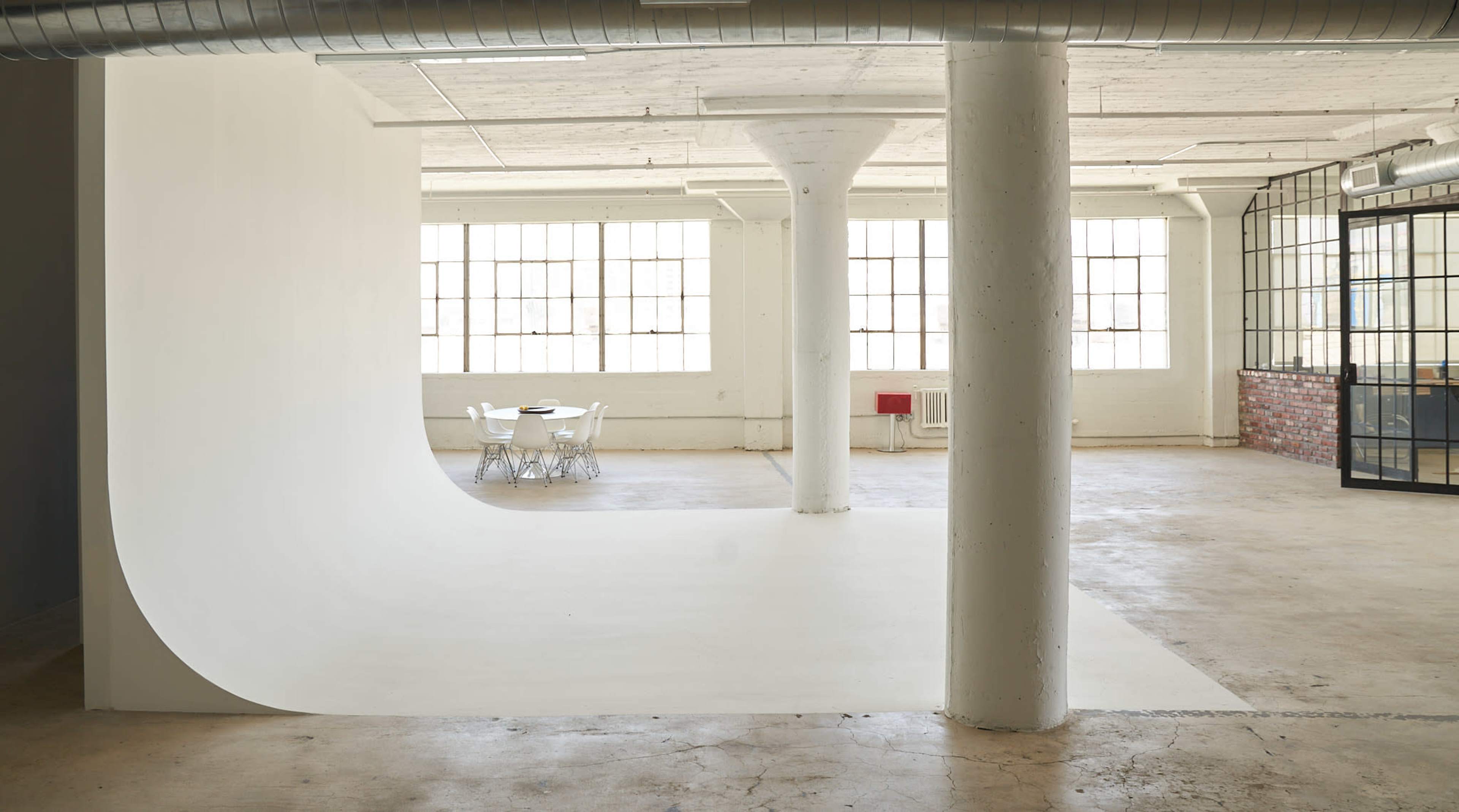 A spacious industrial-style studio with a curved white wall, large windows, and a table set with chairs on a concrete floor.