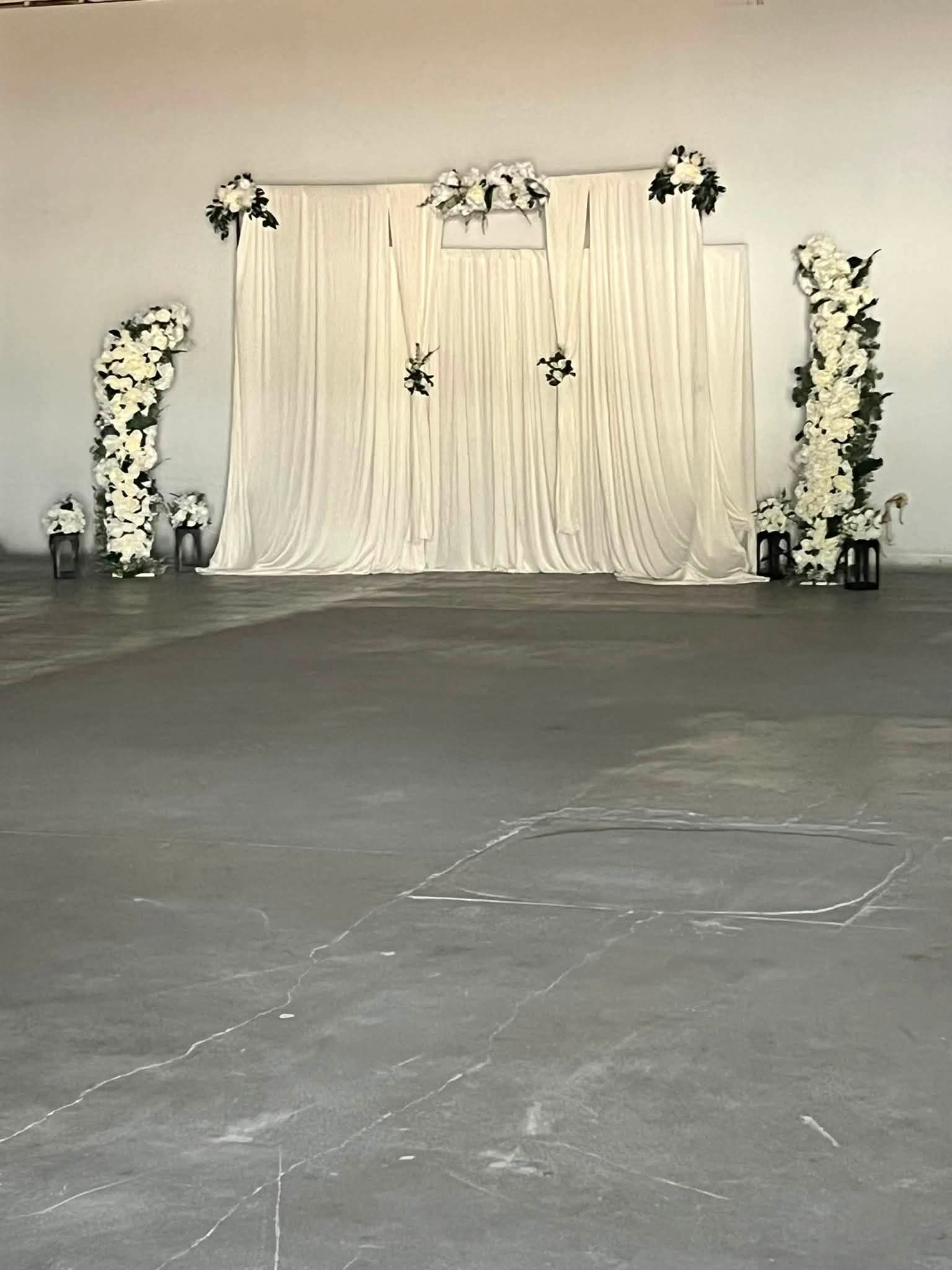 The image shows a backdrop draped with white fabric, flanked by floral arrangements, set up in a spacious, empty interior.