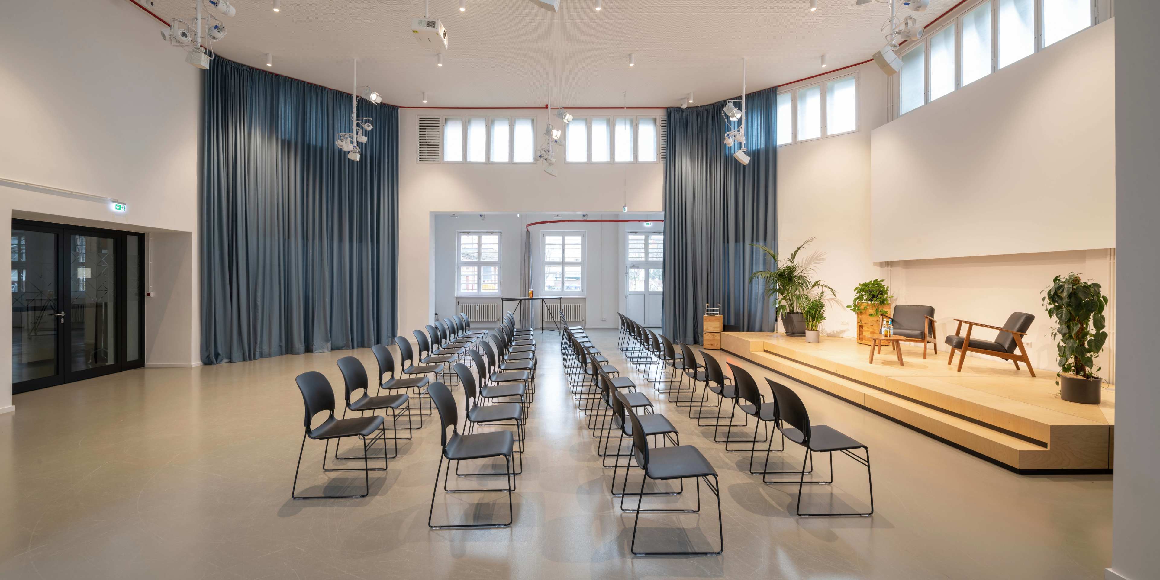 The image shows a spacious auditorium set up with rows of black chairs facing a raised wooden stage, adorned with plants and minimalist decor.