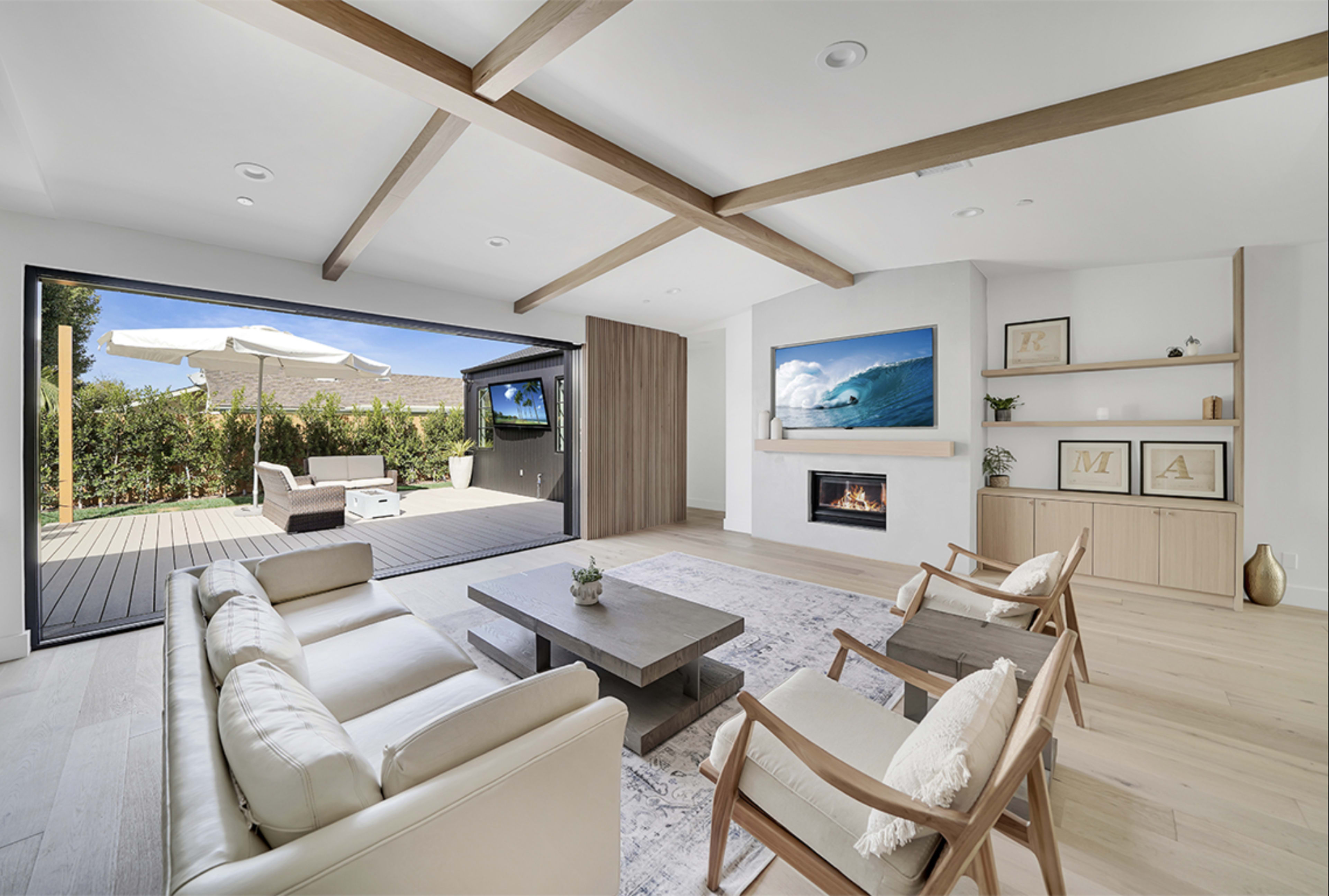 A modern living room features a light-colored sofa, two wooden chairs, a coffee table, and a fireplace, with large glass doors opening to an outdoor seating area.