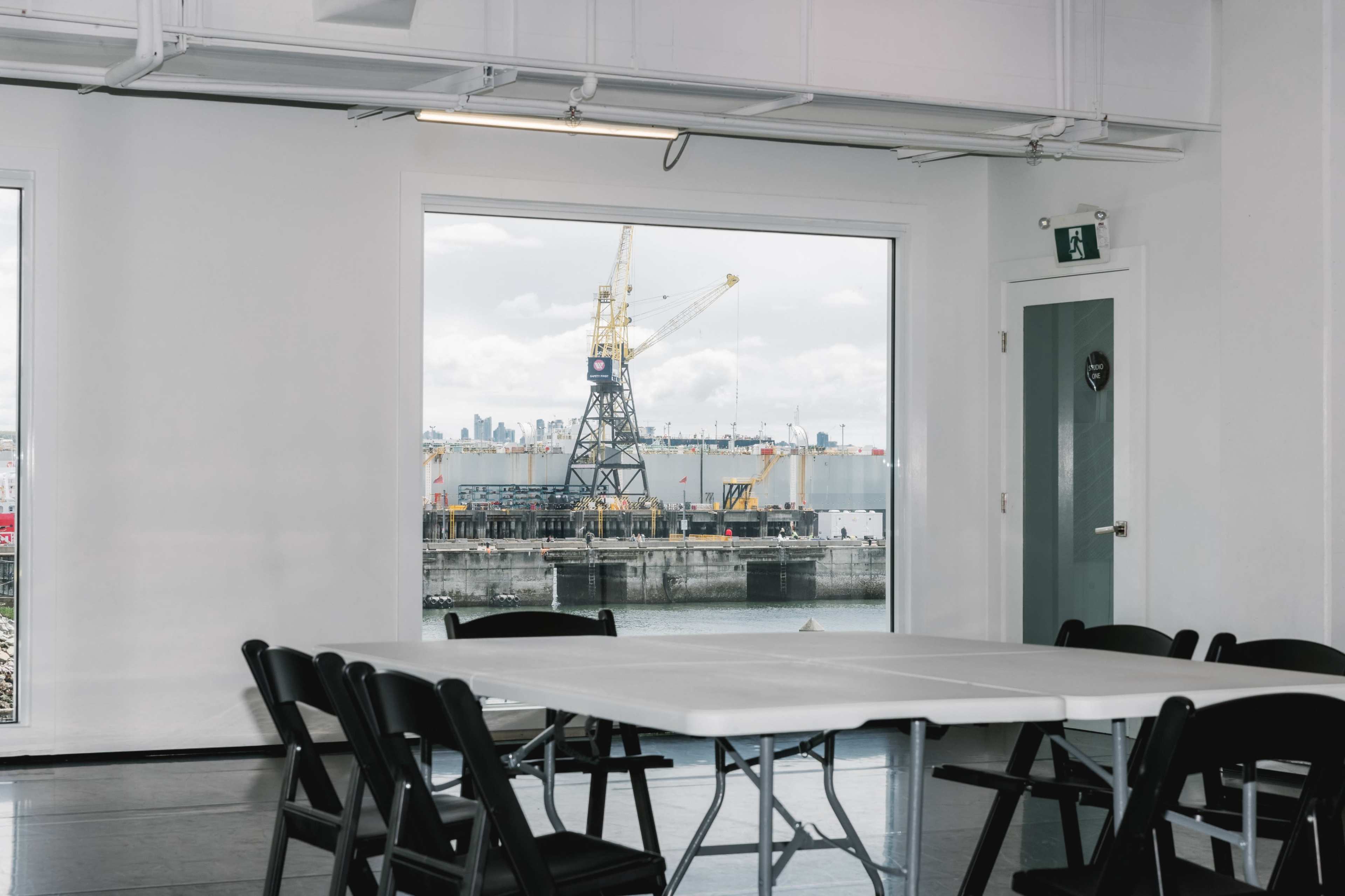 A spacious room with a white interior features a table and black chairs, with a large window overlooking a dock and a crane in the background.