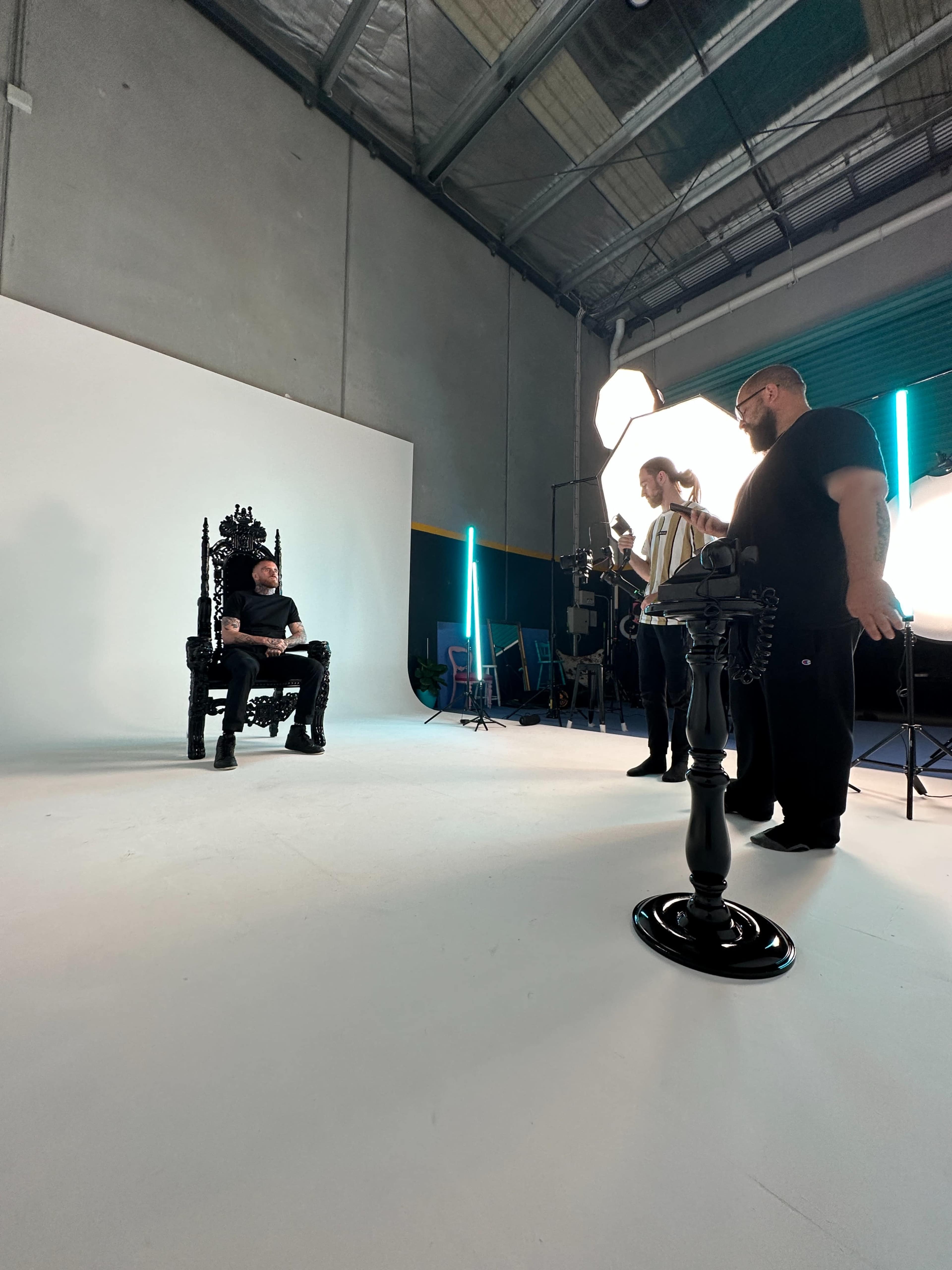 A man sits on an ornate throne in a studio setting while crew members adjust lighting and equipment around him.