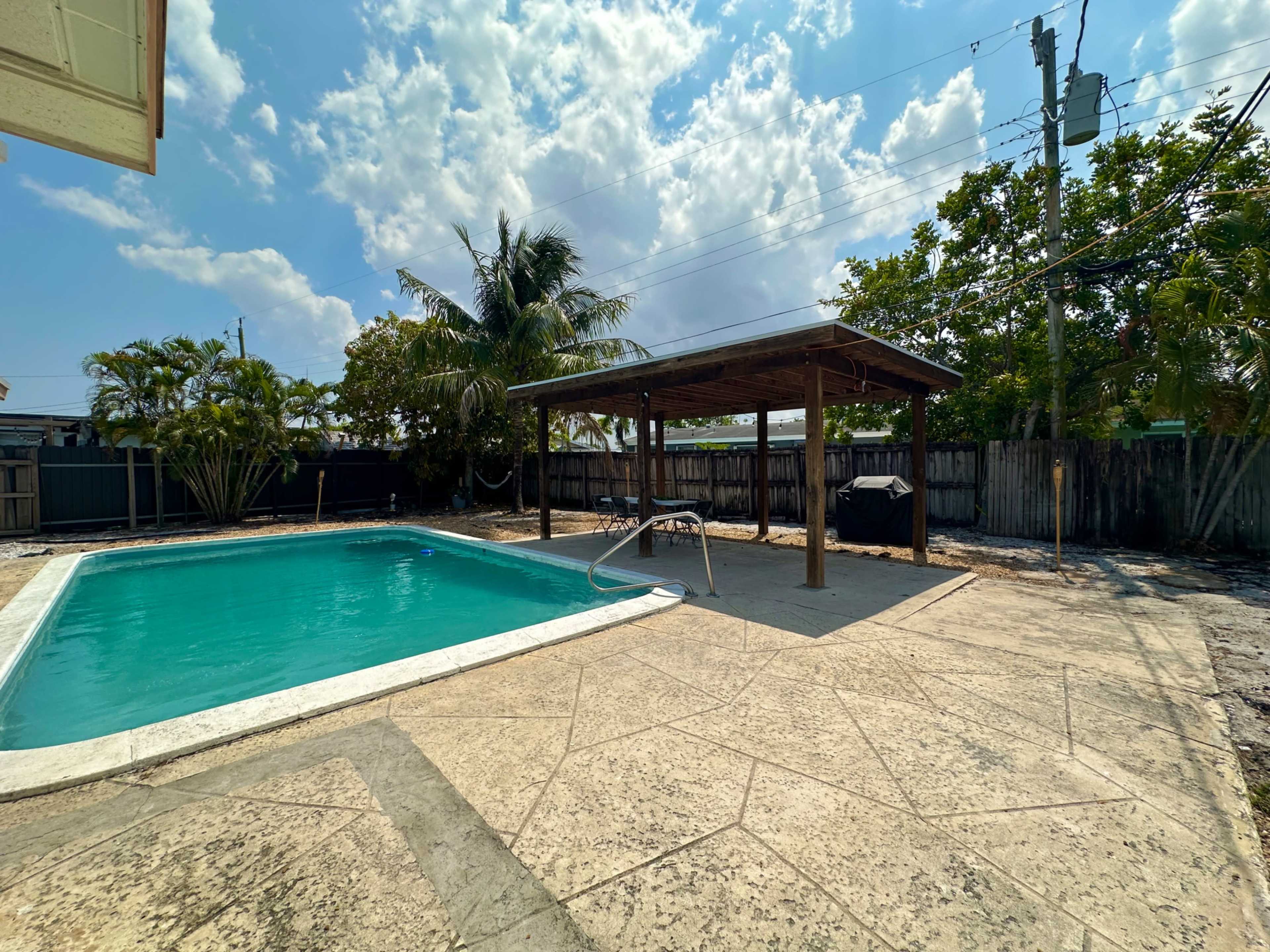 A swimming pool beside a covered patio area, surrounded by palm trees and a fenced yard under a partly cloudy sky.