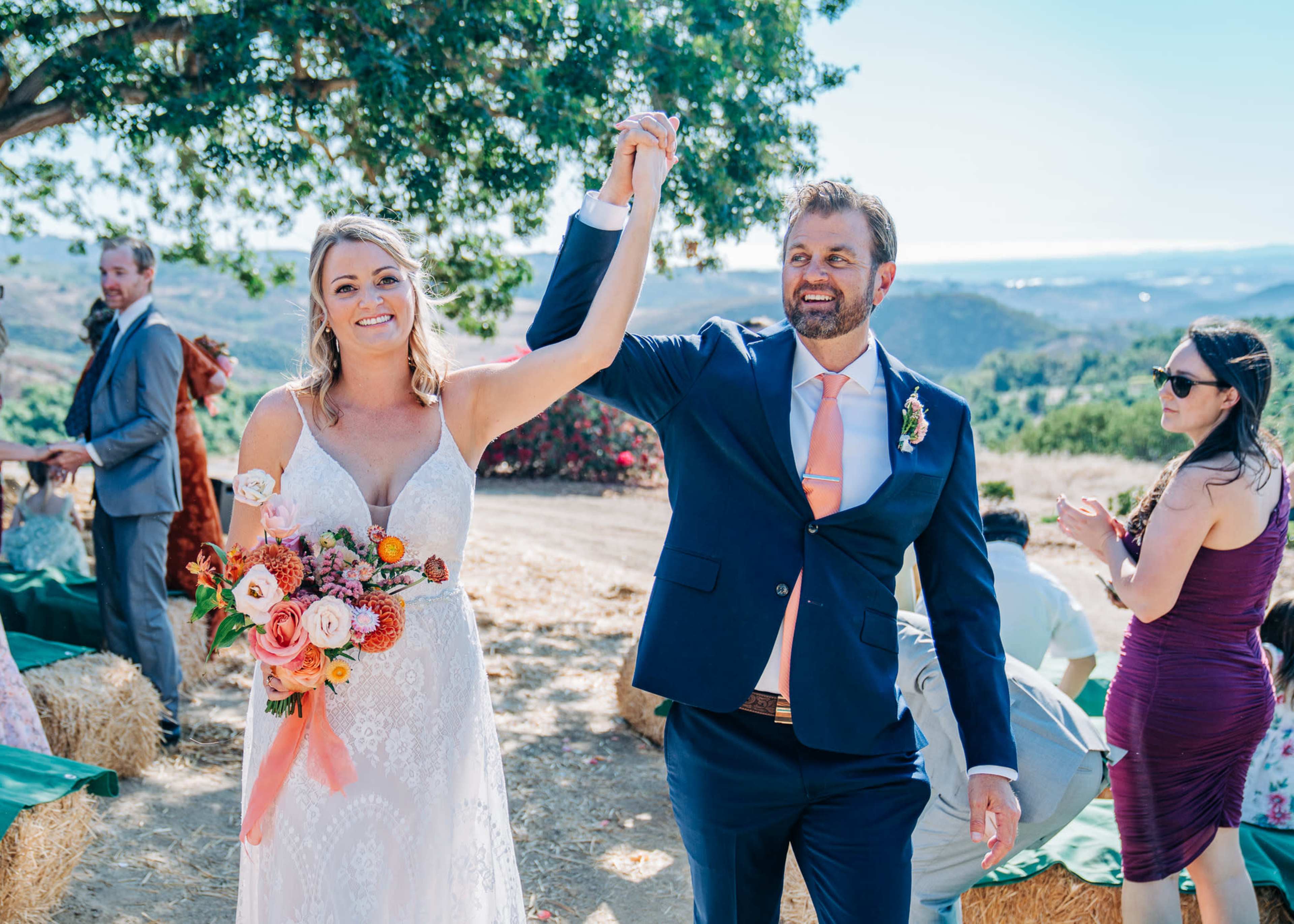 A bride and groom hold hands and celebrate after their wedding ceremony, surrounded by a scenic outdoor setting.