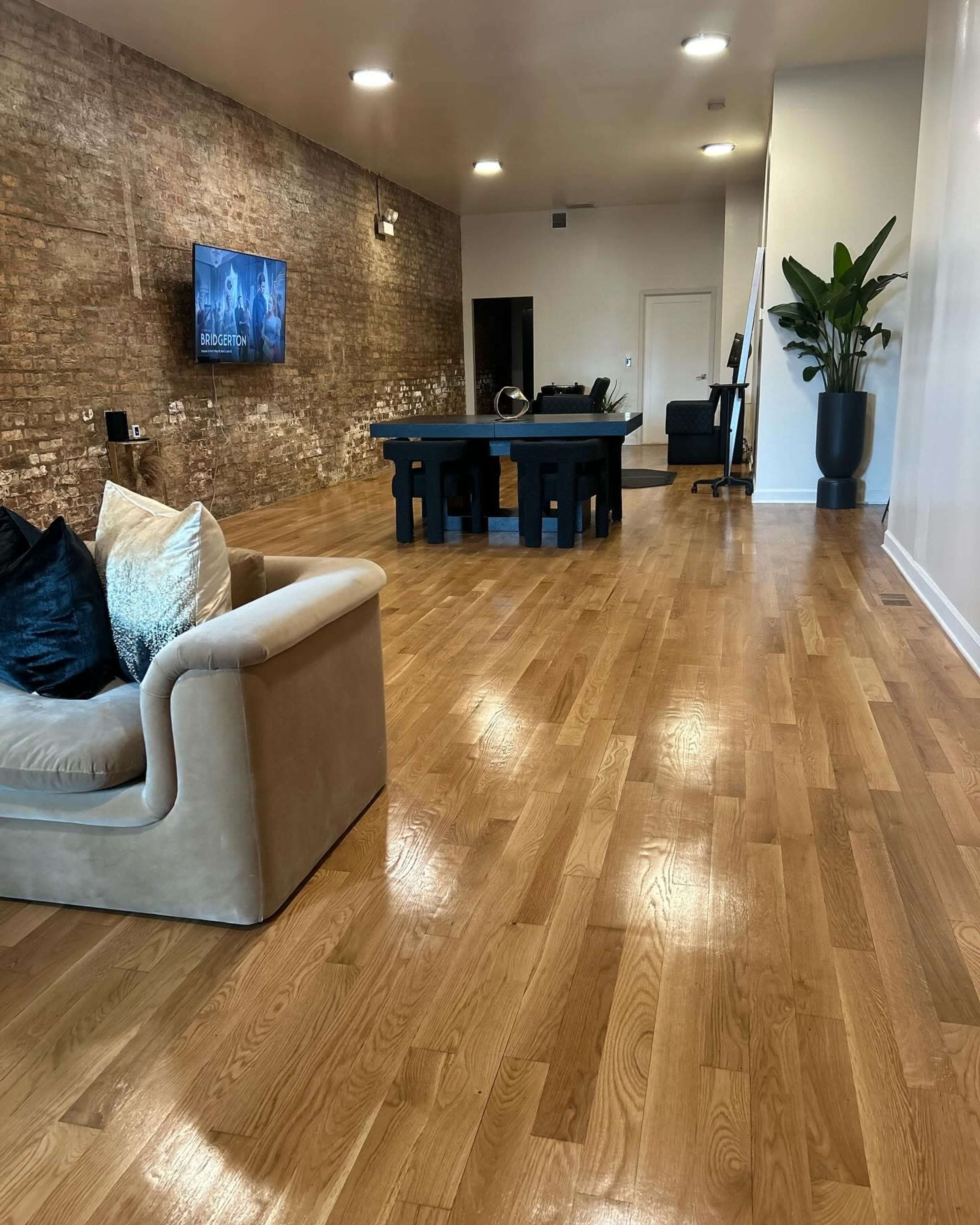 A spacious room with wooden flooring, a sectional sofa, a large dining table, and a potted plant, along with a wall-mounted TV and an office chair in the background.
