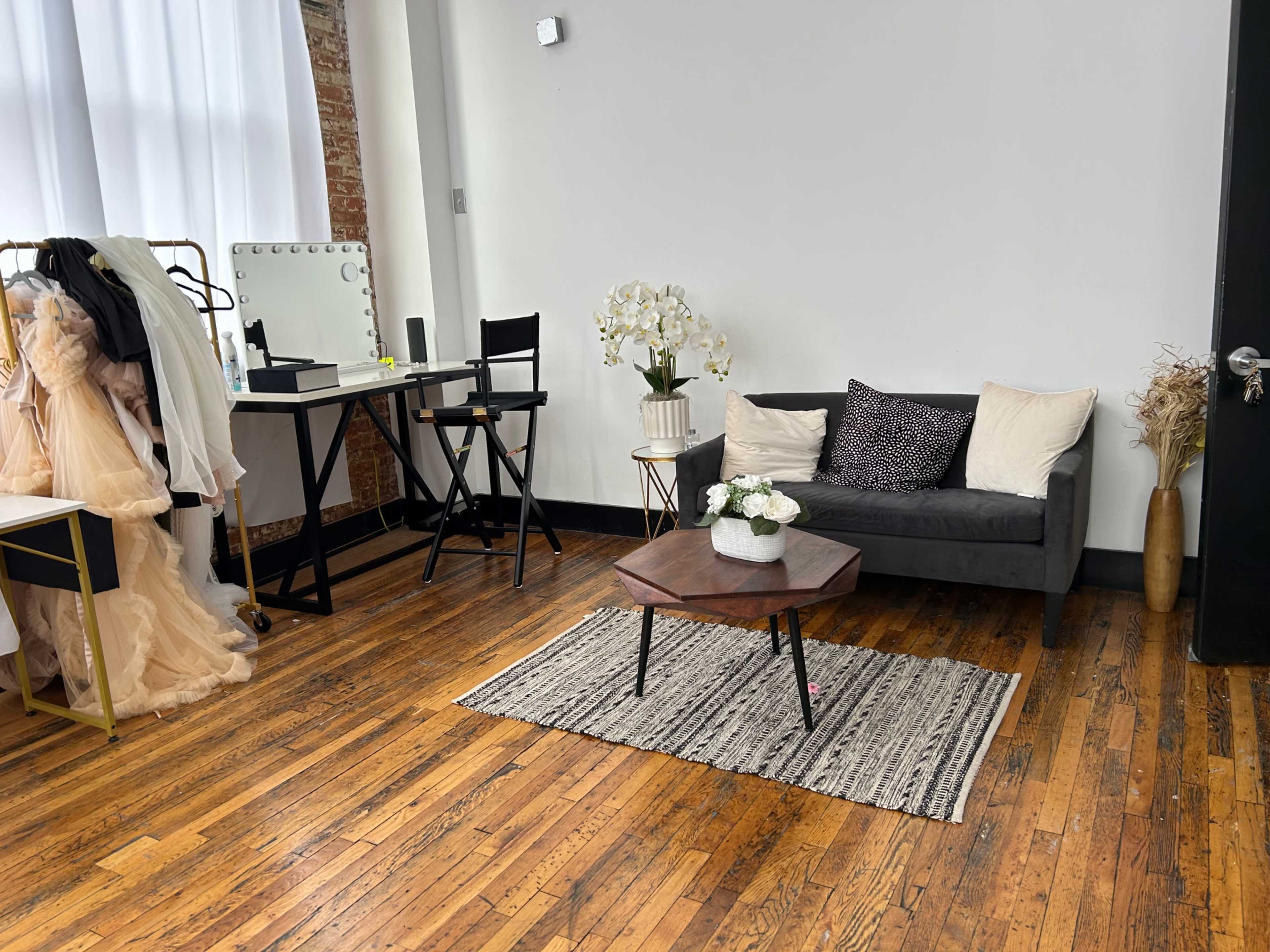 A stylish room with a gray sofa, a wooden coffee table, a makeup station, and a rack displaying dresses, all set on a wooden floor.