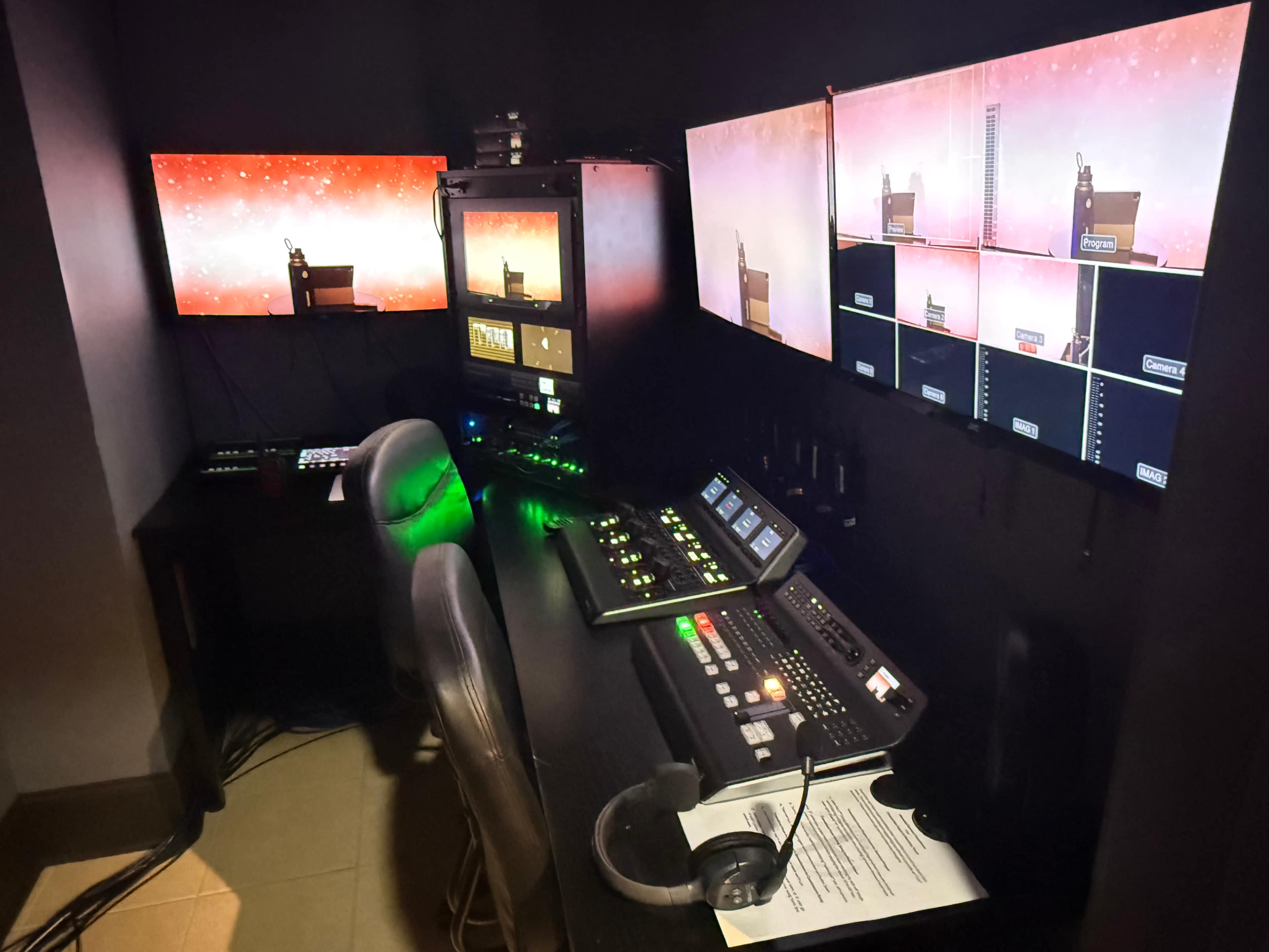 The image shows a control room with multiple monitors and control panels arranged on a dark wall.