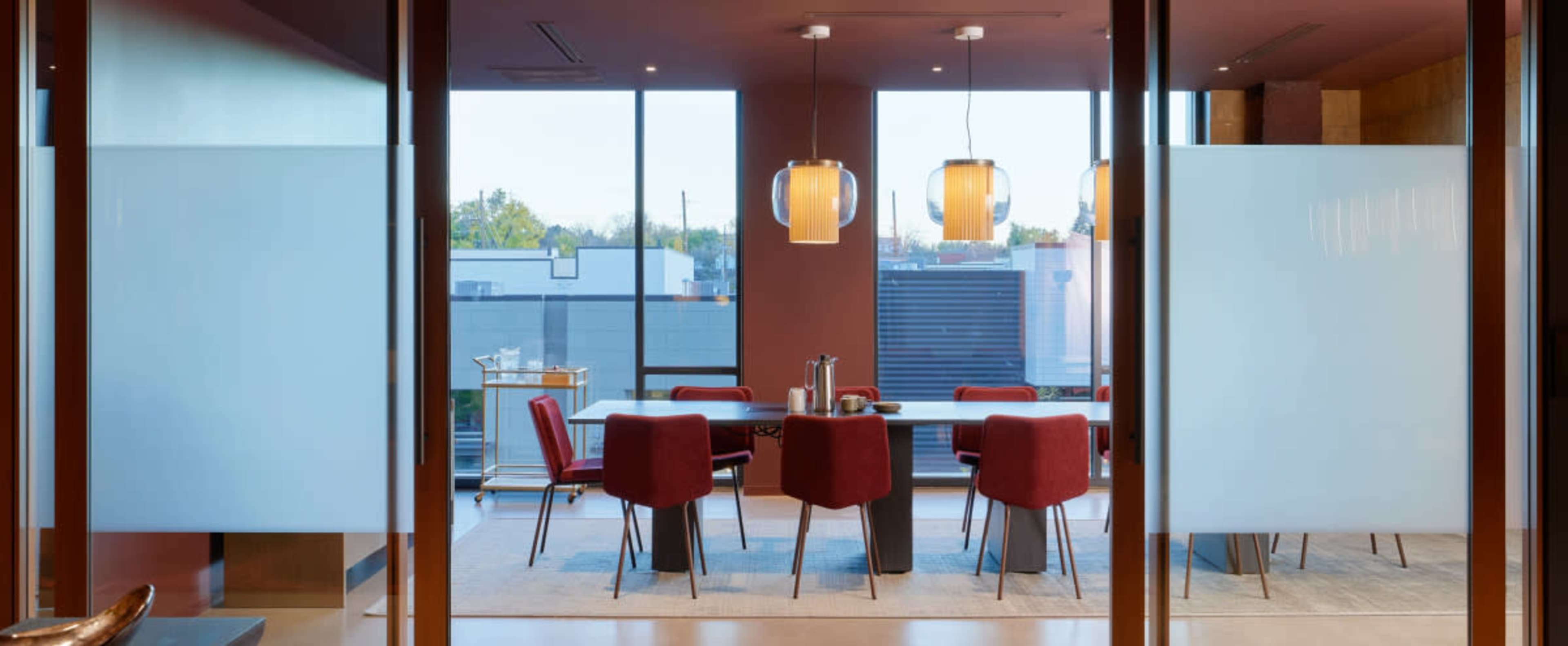 A modern dining area features a large table surrounded by red chairs, with pendant lighting and large windows providing natural light.