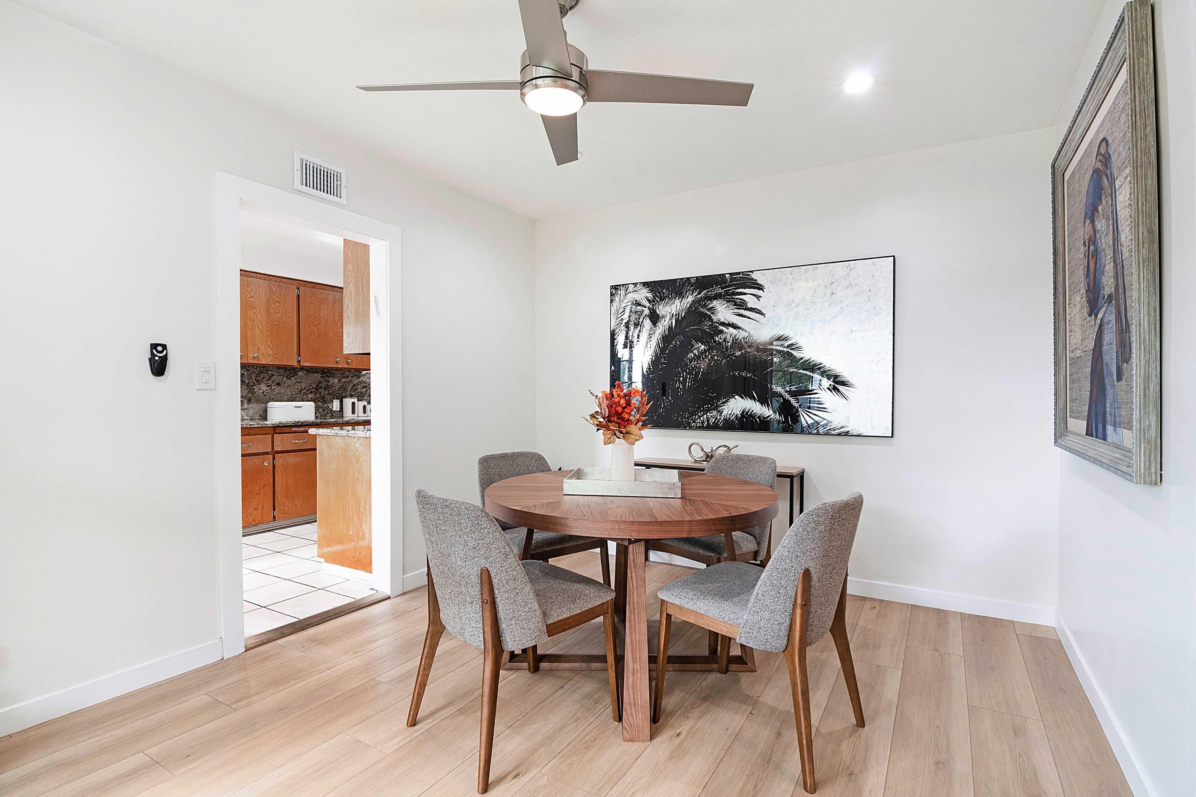 A dining area features a round wooden table surrounded by four grey upholstered chairs, a large wall art piece, and a ceiling fan.