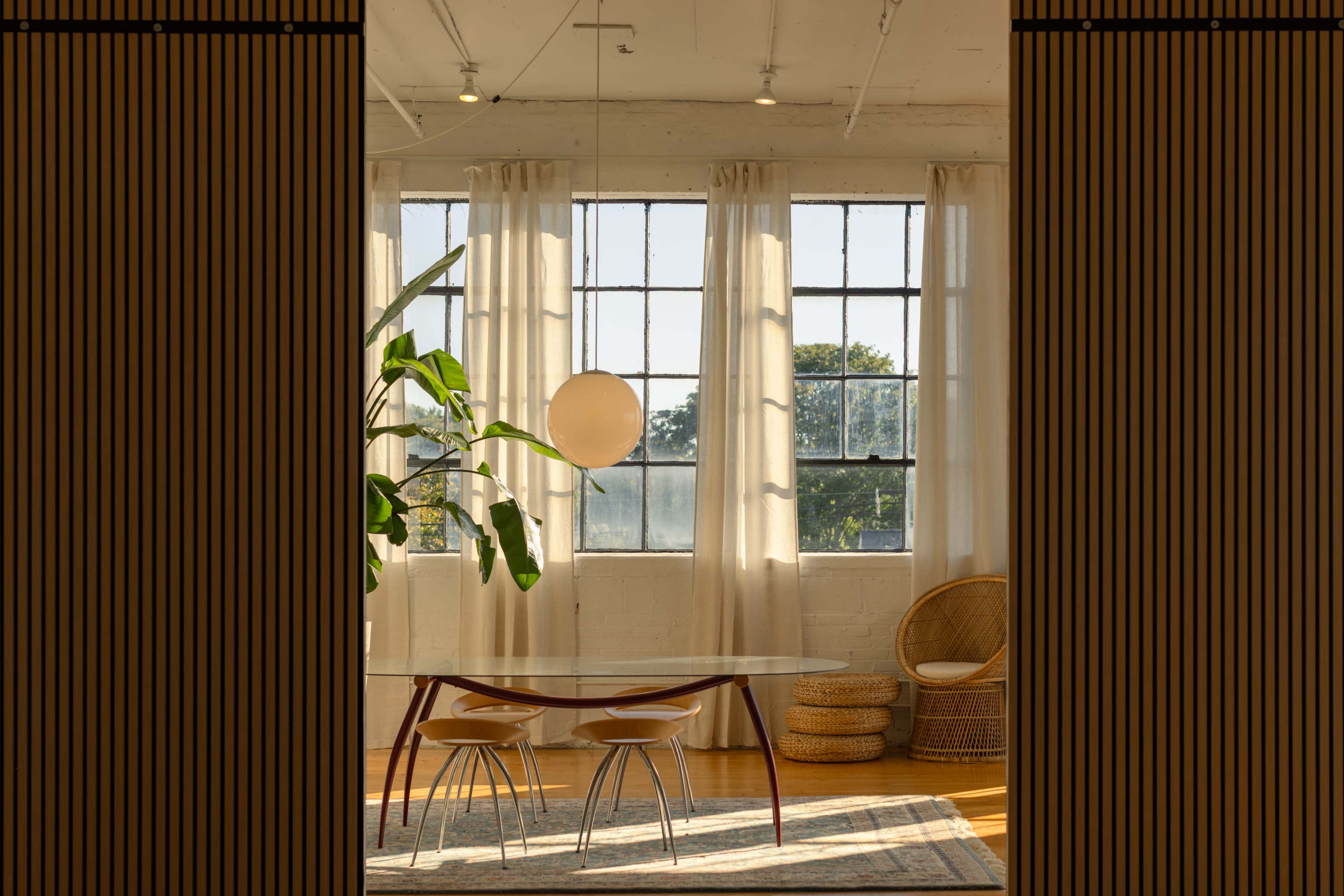 The image shows a bright interior space featuring a table with two chairs, a large plant, and windows with sheer curtains.