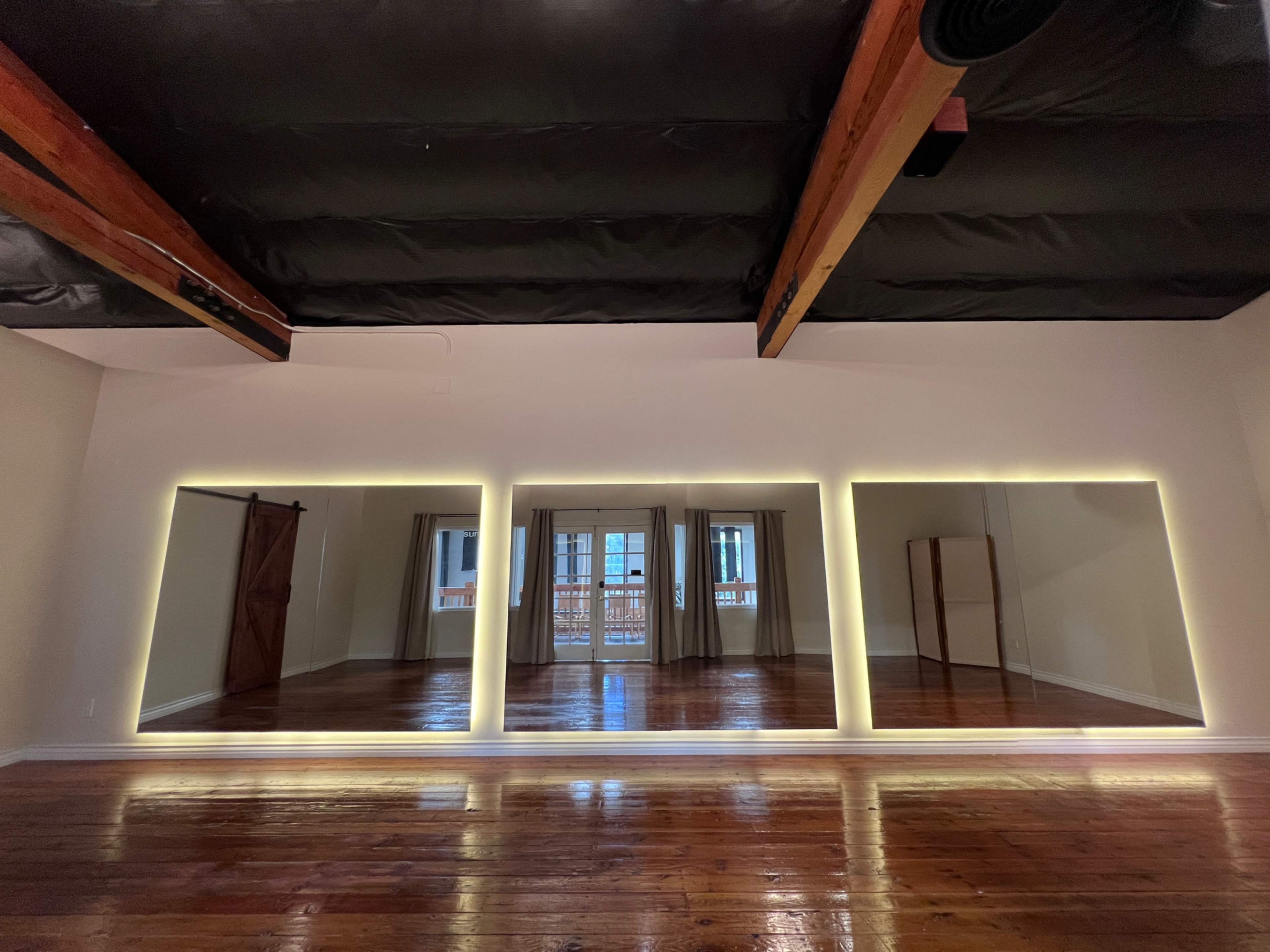 The image shows a spacious room with three large backlit mirrors and hardwood flooring, featuring wooden beams on the ceiling and a doorway leading to a balcony.