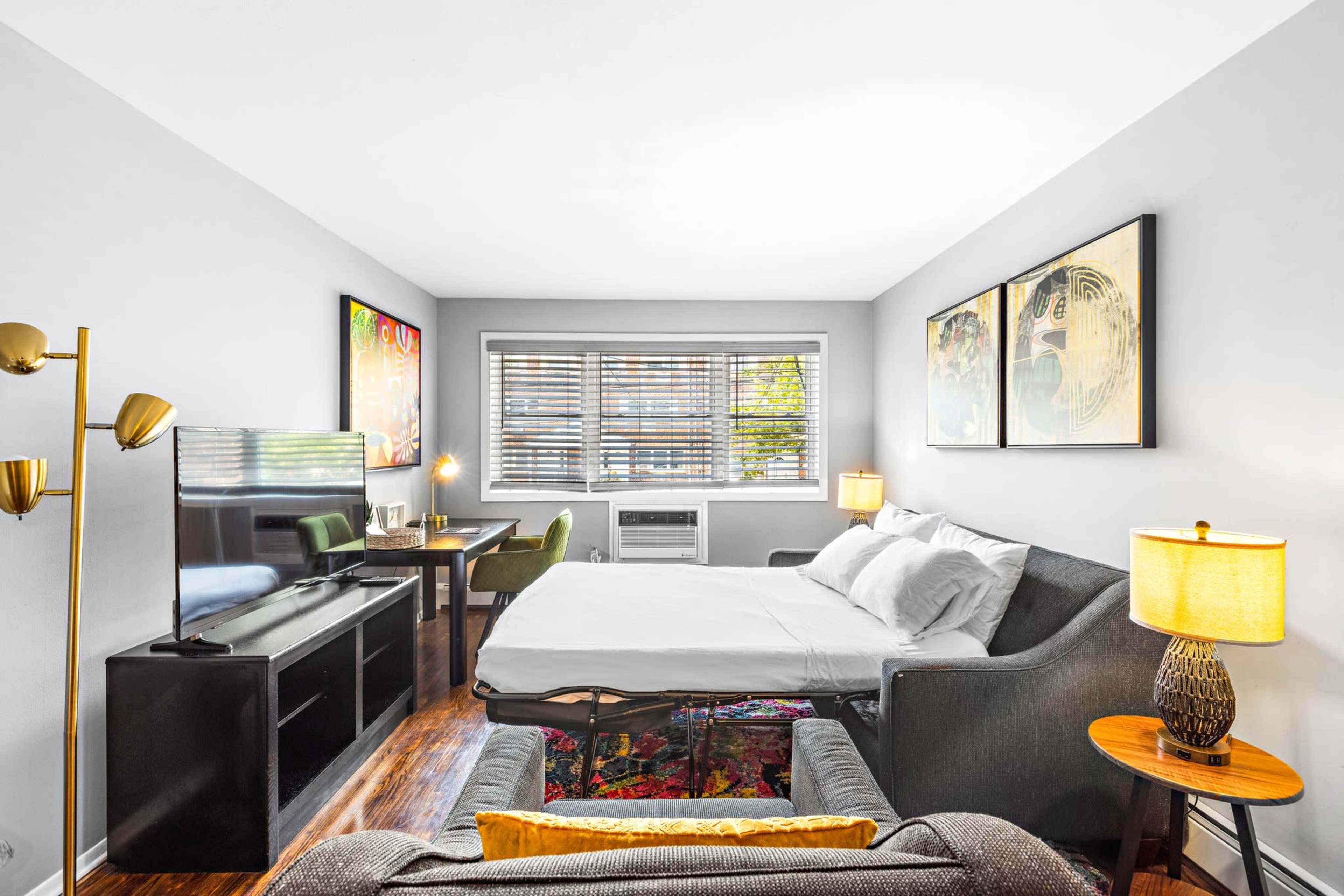 A hotel room featuring a bed, a small desk with a chair, a television, and two lamps, with large windows providing natural light.