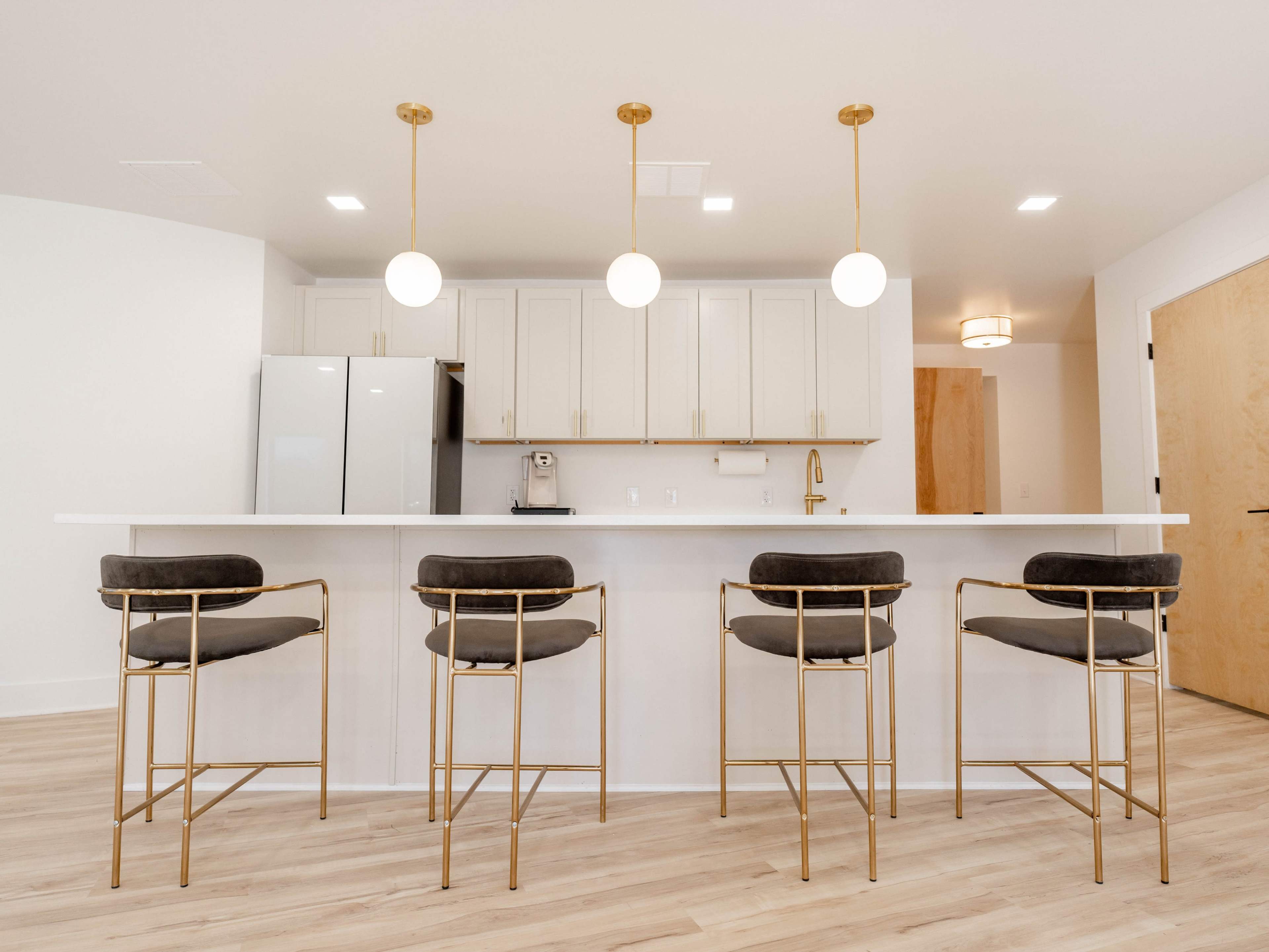 A modern kitchen with a white countertop, four gold-framed bar stools, and pendant lights hanging above.