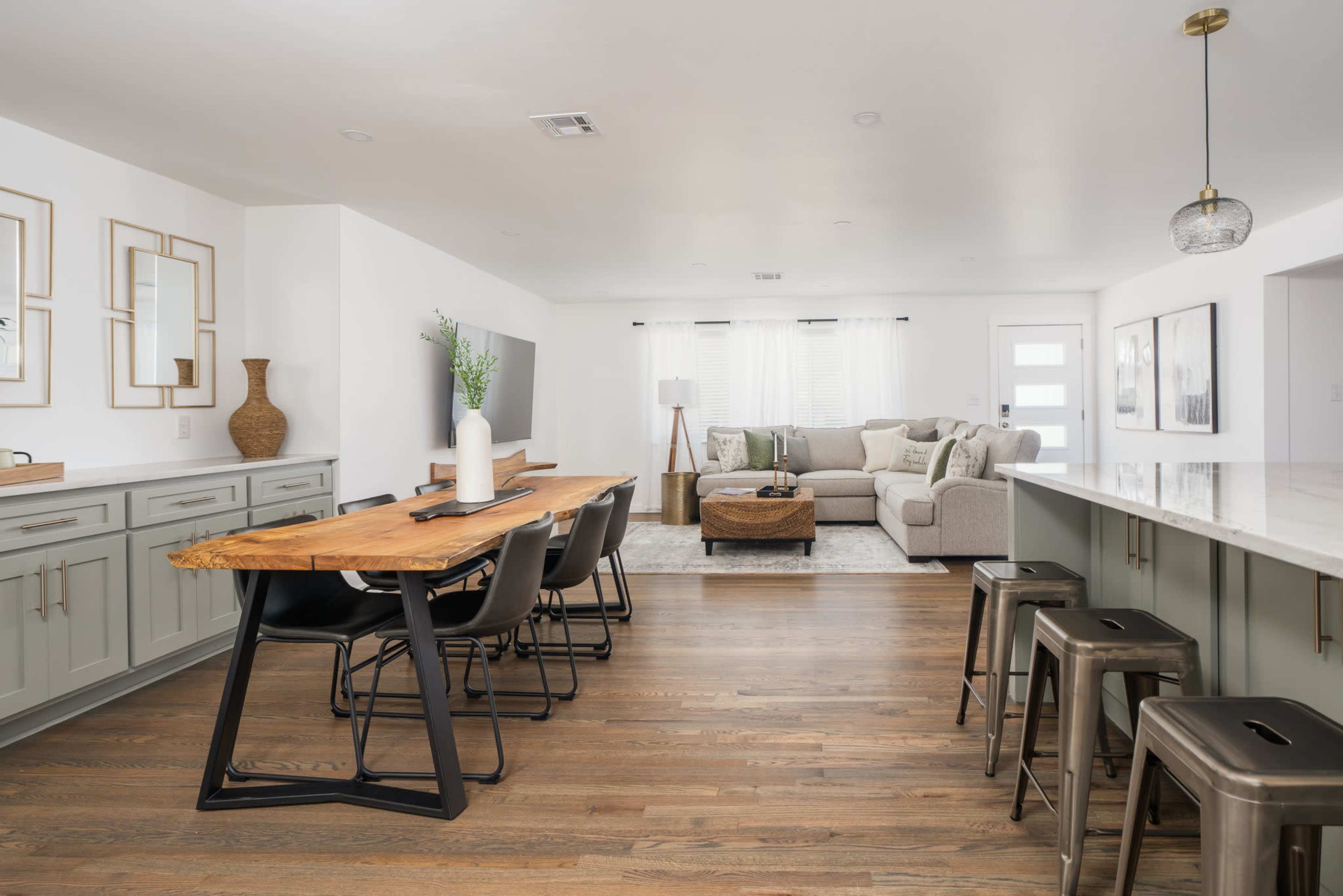 A modern open-concept living space featuring a dining area with a wooden table and black chairs, a cozy seating area with a light gray sofa, and a kitchen with bar stools.