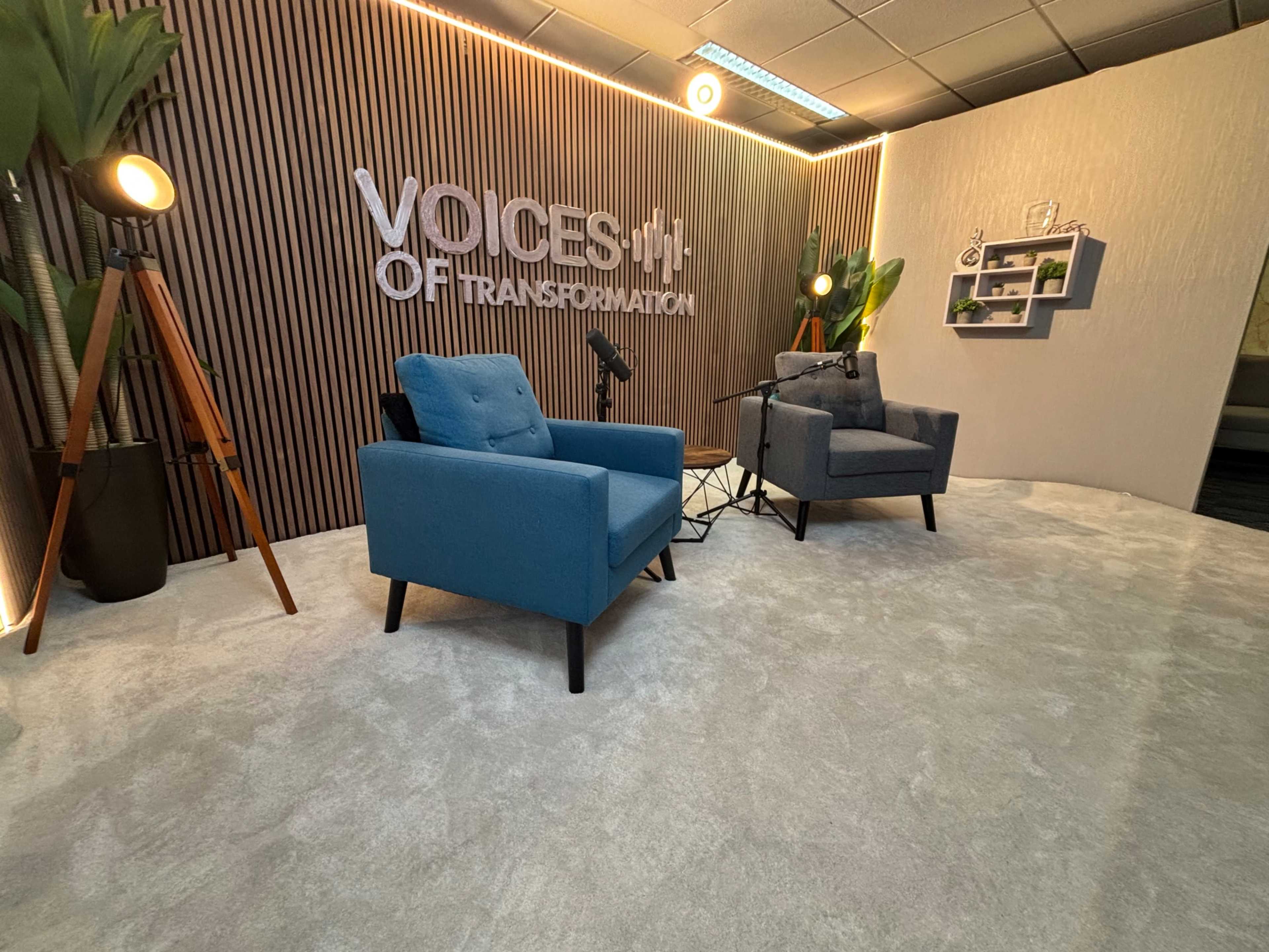 The image shows a modern interview or podcast setup featuring two blue chairs, a small round table, and a backdrop with the words "VOICES OF TRANSFORMATION" on a wooden panel.