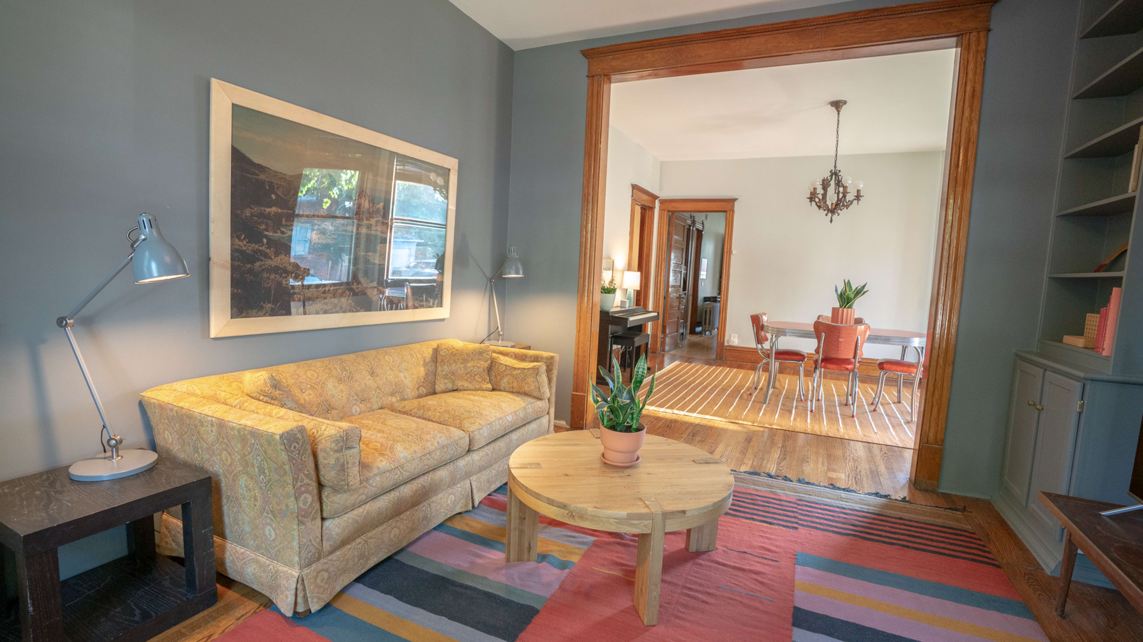 A living room with a patterned sofa, a round wooden coffee table, and a view into a dining area featuring a chandelier and additional seating.