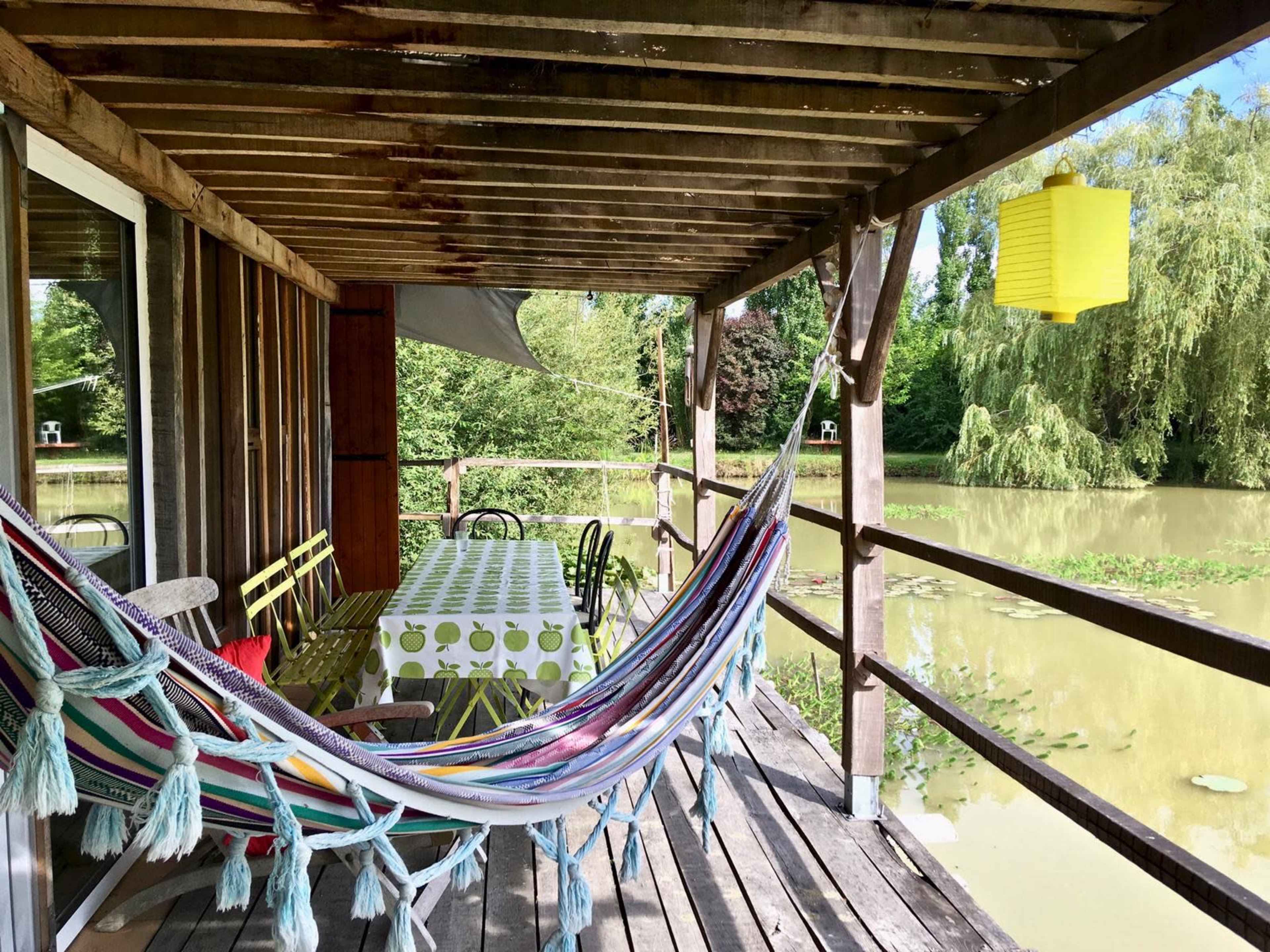 A wooden porch overlooks a pond, featuring a striped hammock, a table covered with a patterned cloth, and a yellow lantern hanging from the ceiling.