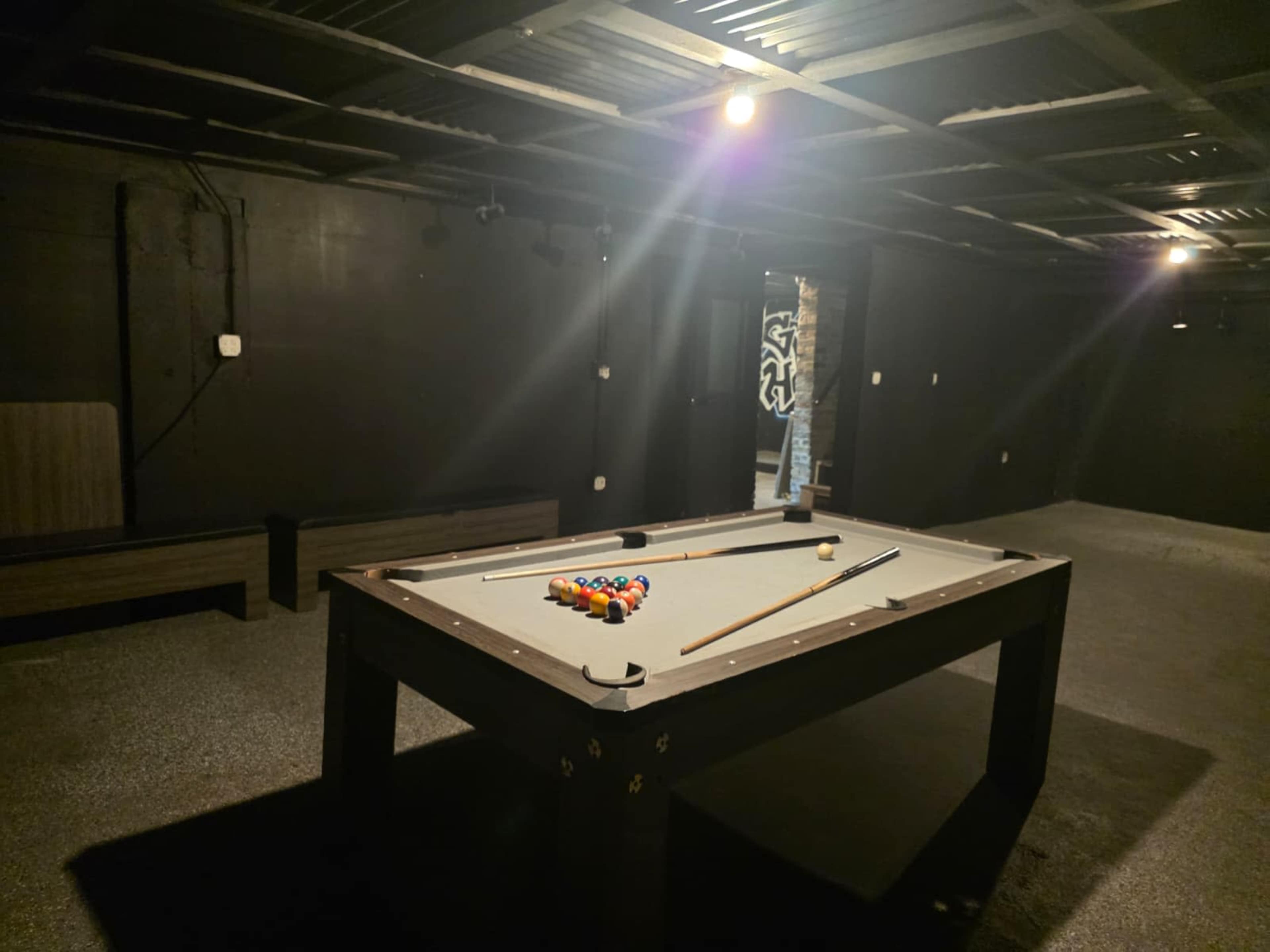 The image shows a pool table with a set of colored balls racked on it, surrounded by dimly lit black walls in an indoor space.