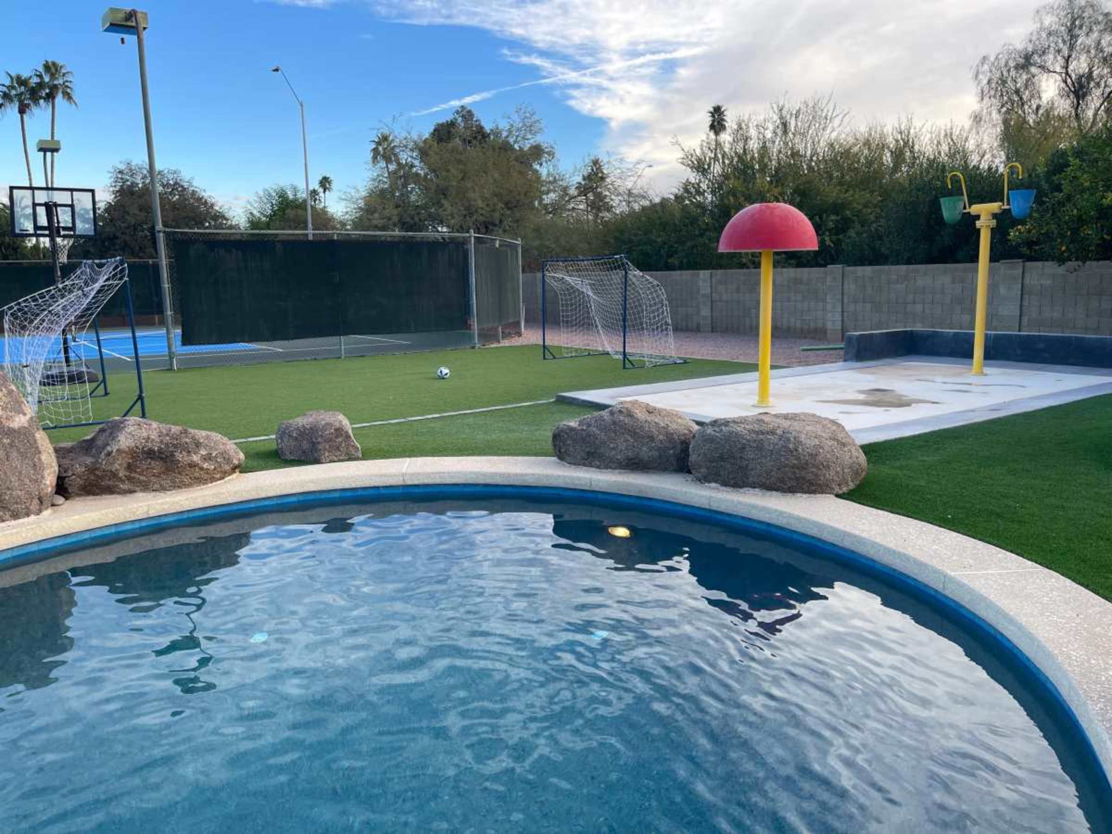 The image shows a backyard featuring a swimming pool, a water play structure, a soccer goal, and a grassy area with a basketball hoop in the background.