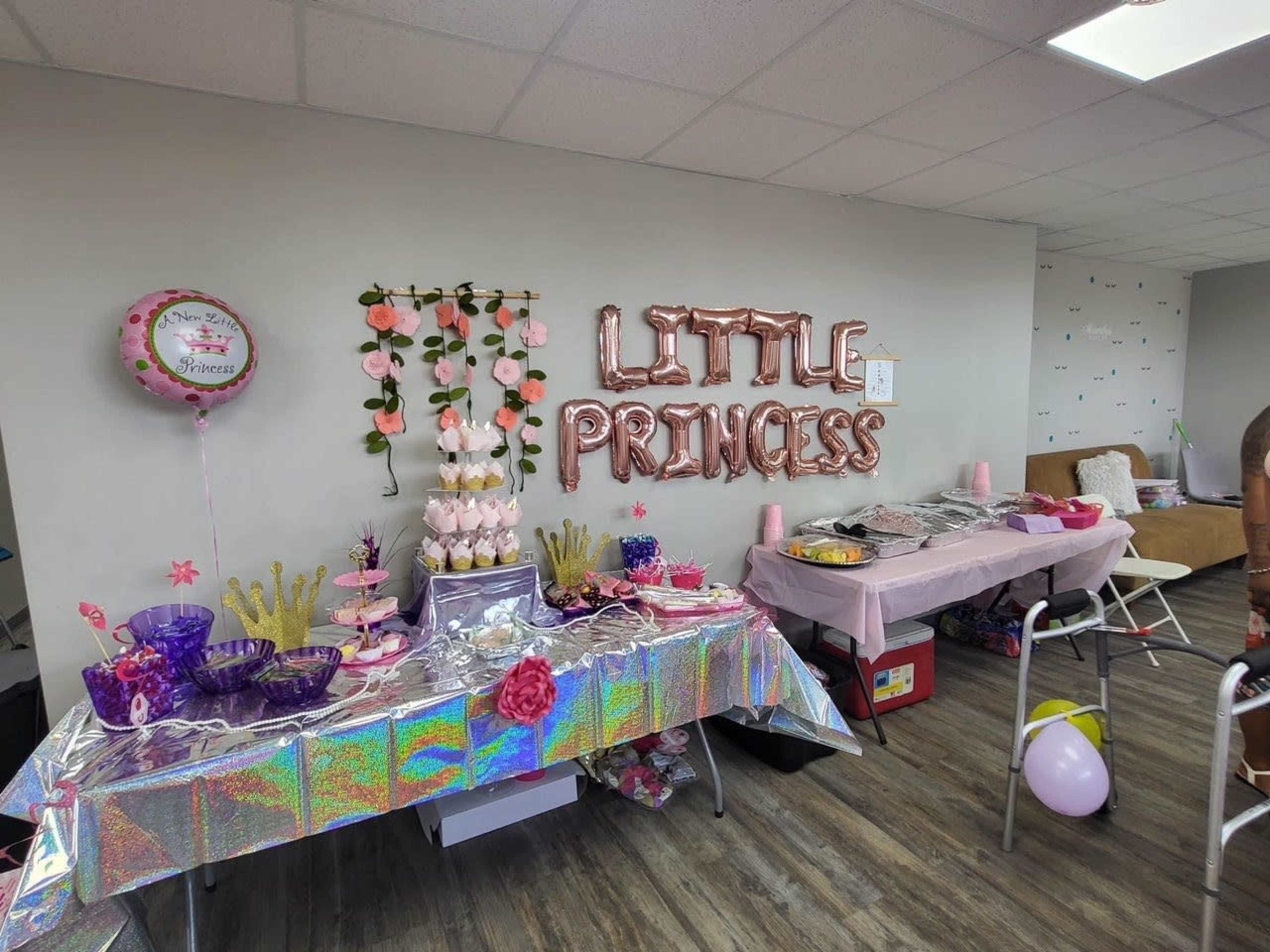 A party setup with a table featuring a pink tablecloth, a tiered cupcake stand, and decorations that read "LITTLE PRINCESS" along the wall.