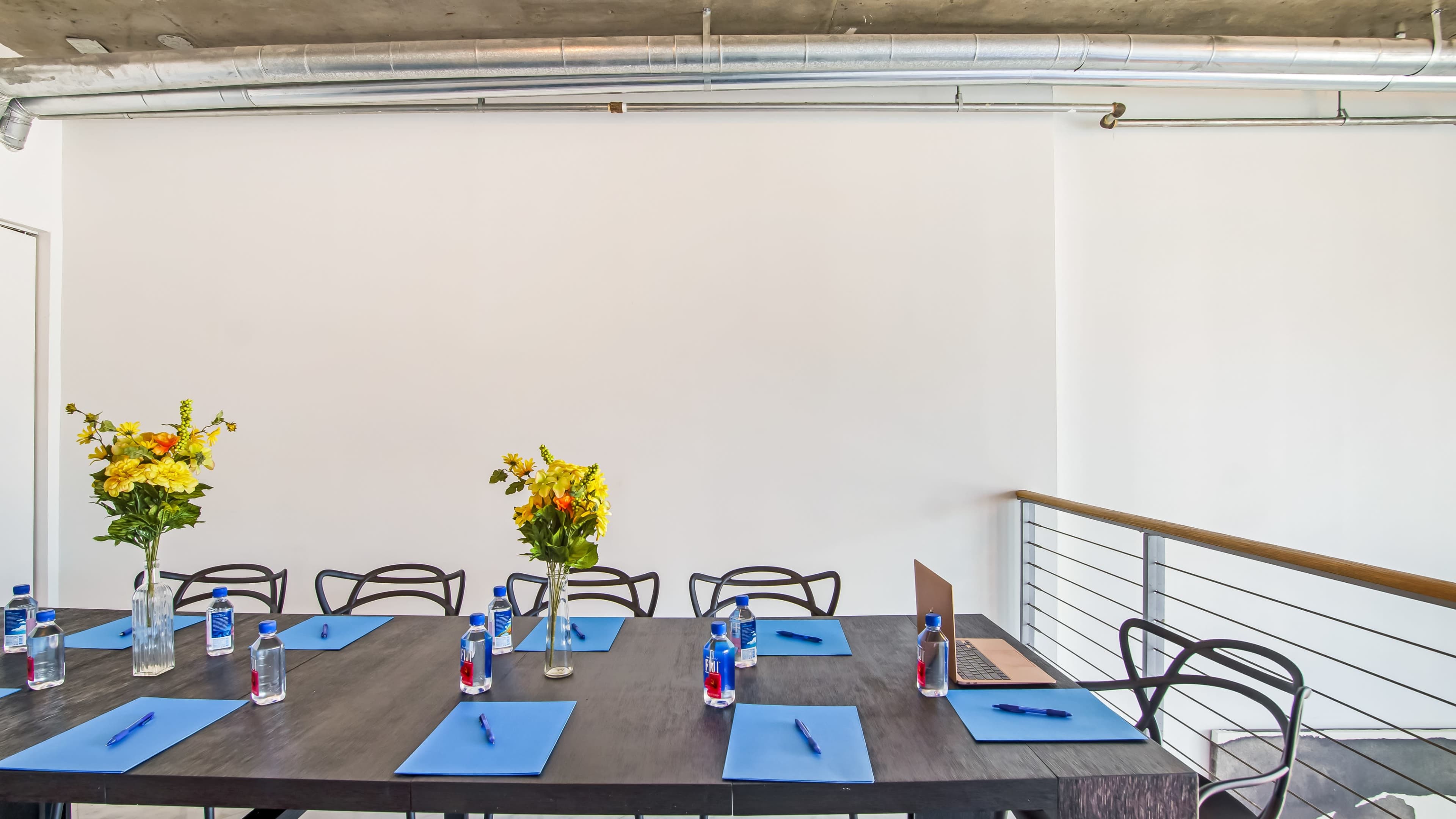 A conference room table is set with blue notepads and pens, featuring two vases of flowers and bottled water.