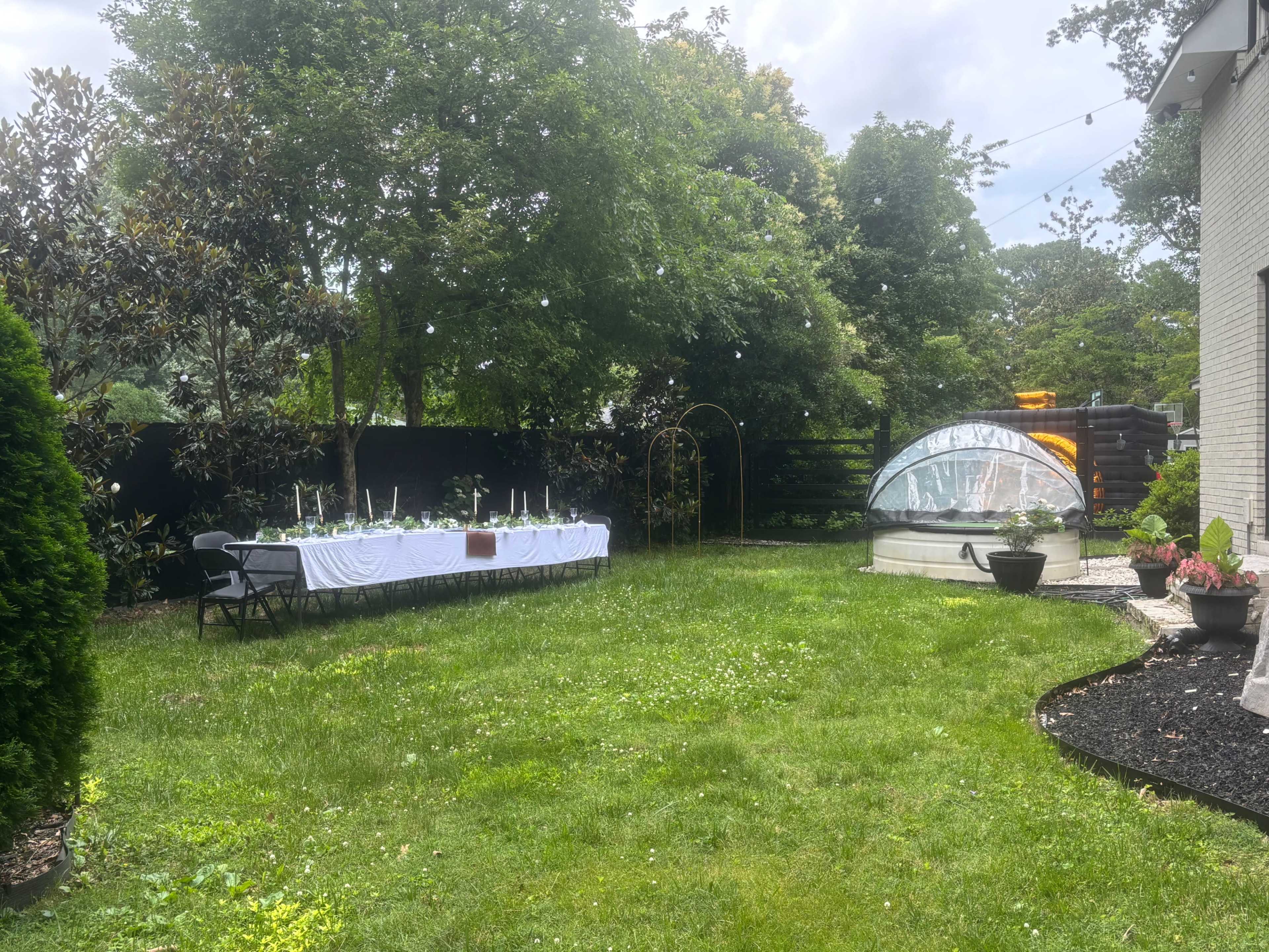 A long table is set up under trees in a grassy backyard, surrounded by decorative plants and a clear dome structure.