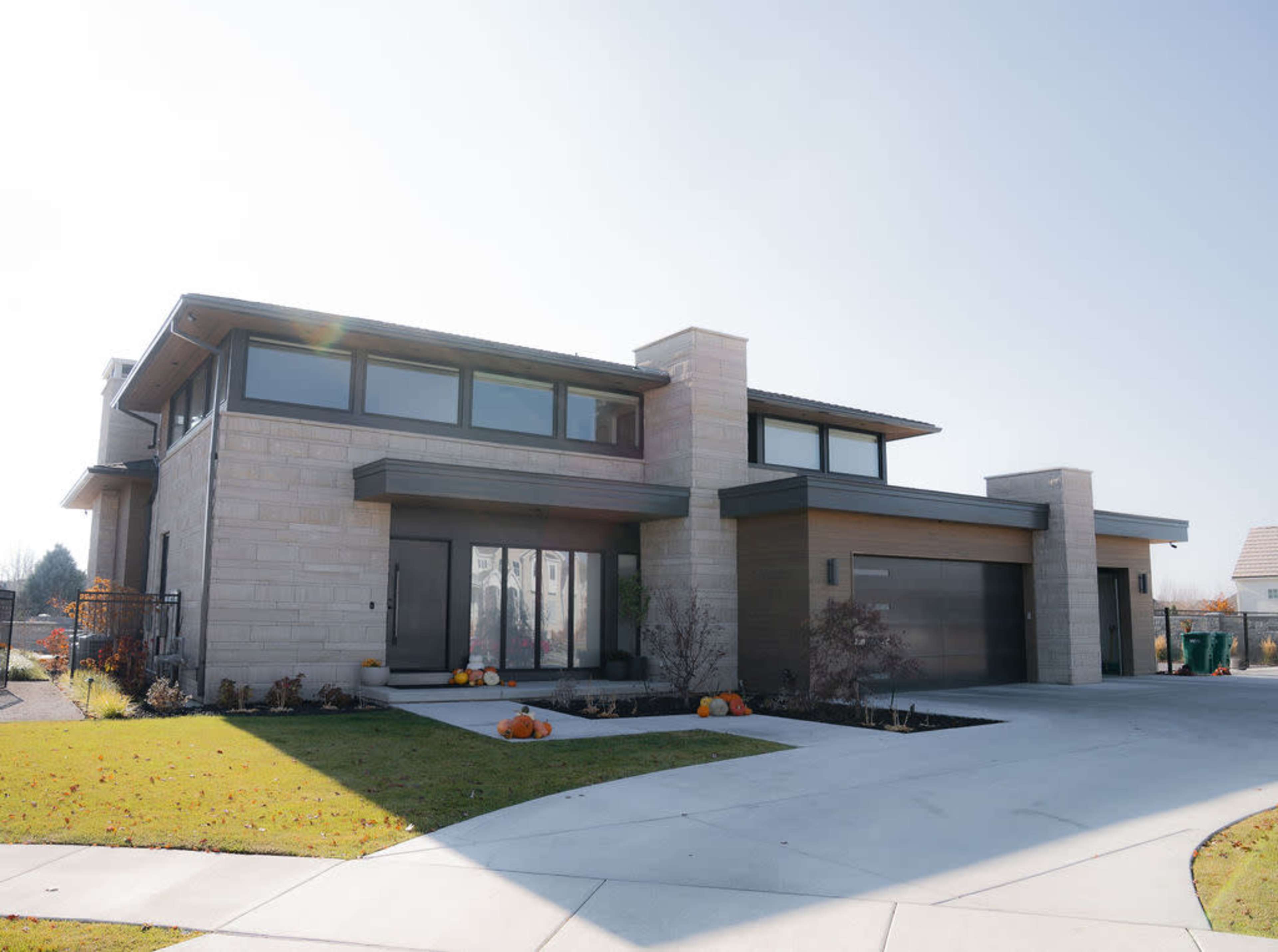 A modern two-story house with large windows and a stone facade features a spacious front yard with pumpkins and a circular driveway.