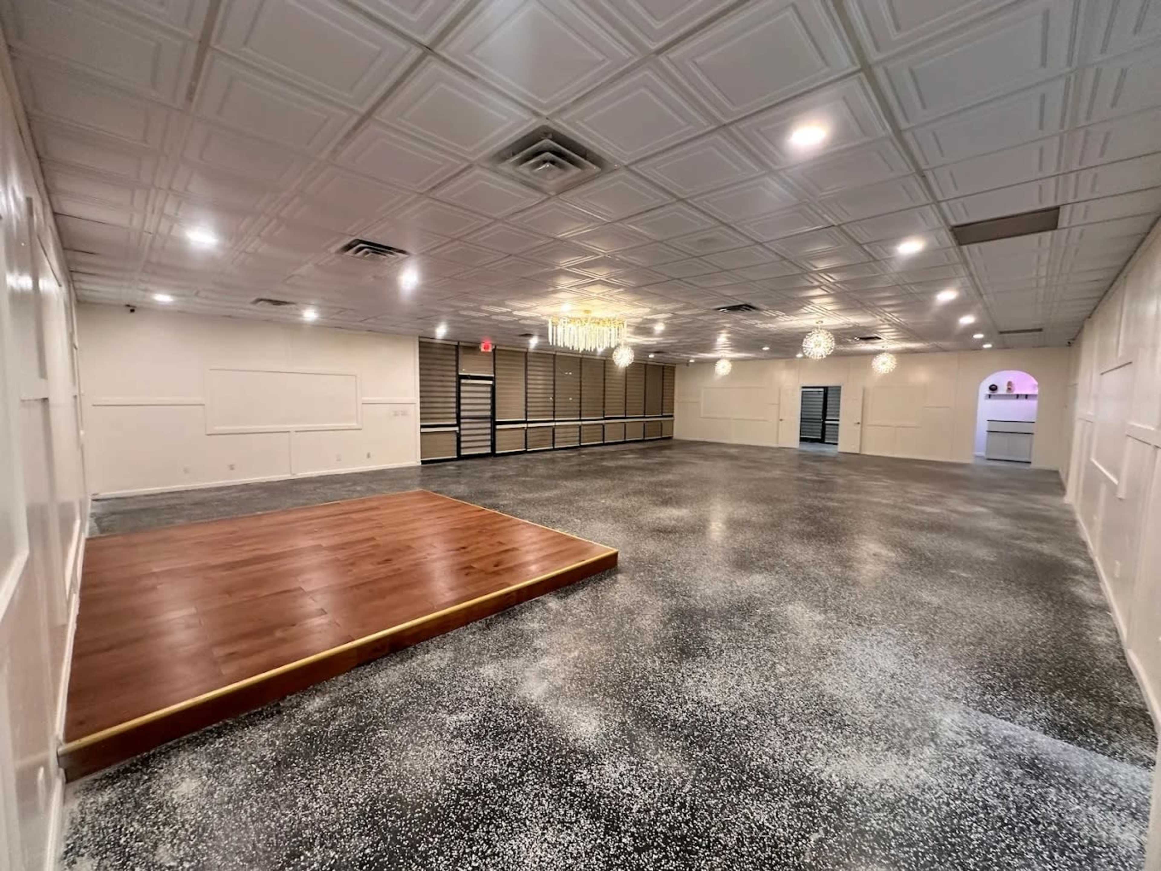 The image shows a spacious, empty room with a raised wooden stage at one end, a textured ceiling, and modern lighting fixtures.