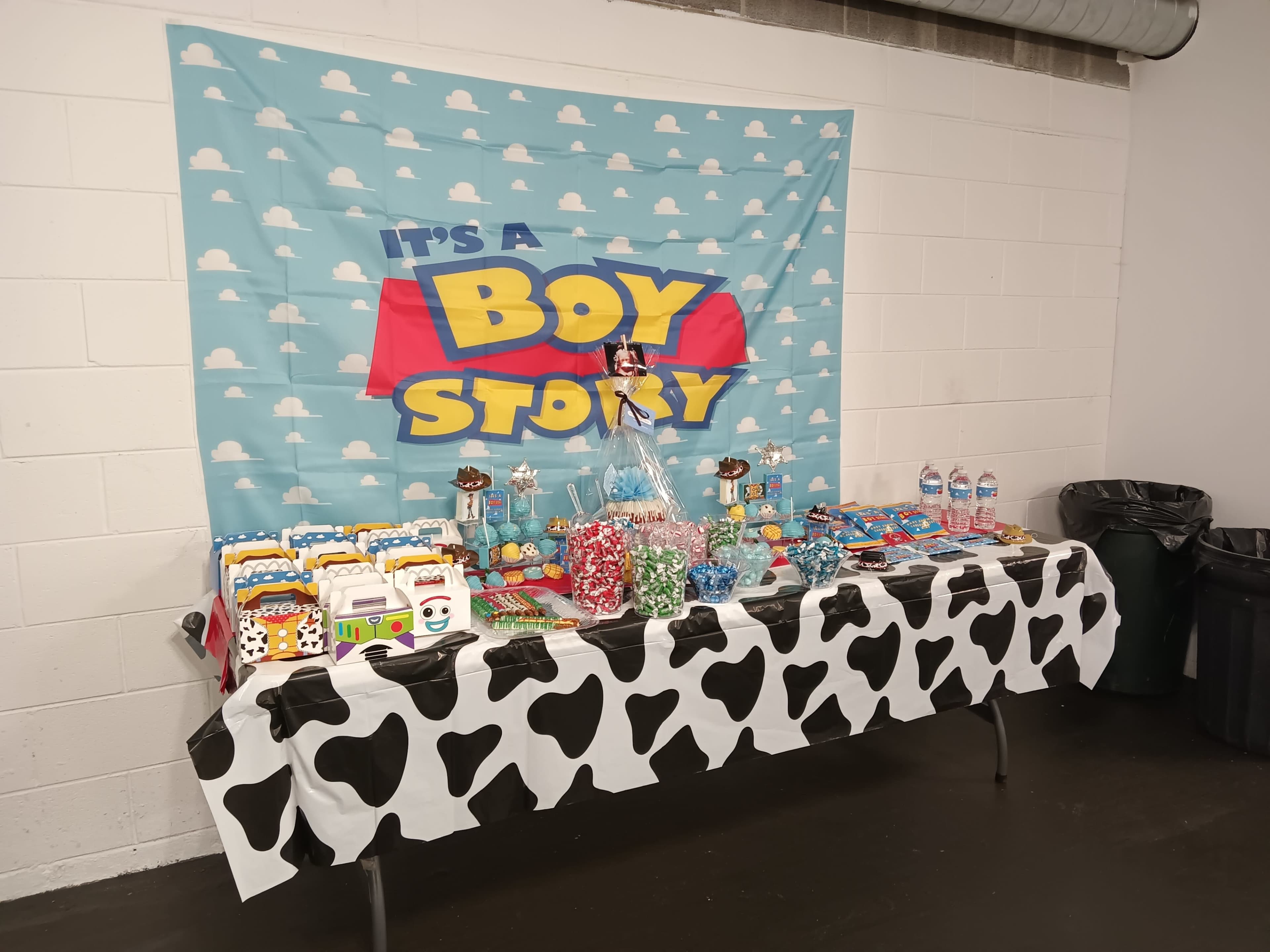 A table is decorated with a cow-print tablecloth and a "It's a Boy Story" backdrop, displaying various colorful candies and snacks in themed containers.