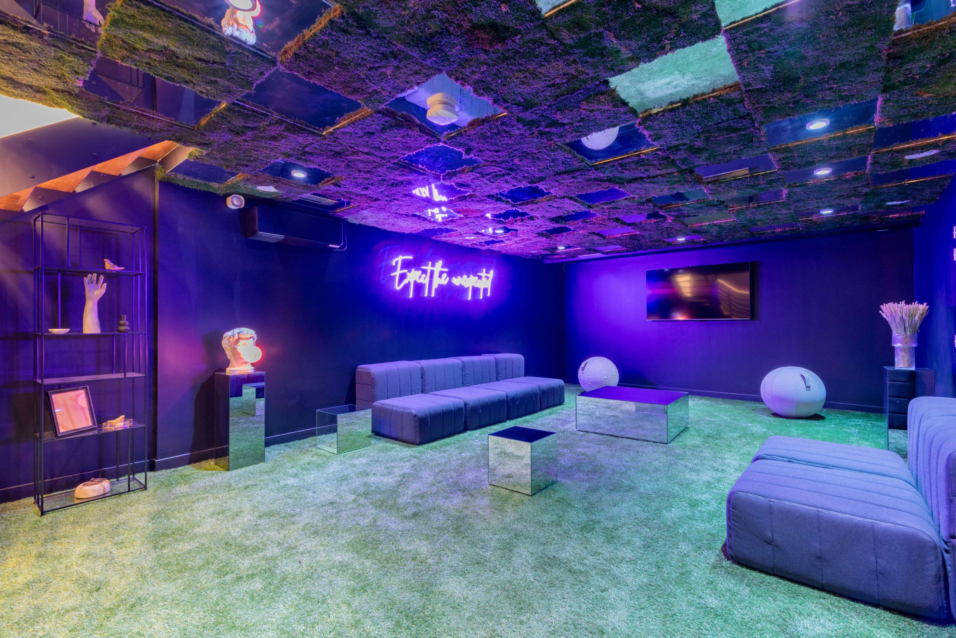 The image shows a modern lounge with a grass-like carpet, mirrored furniture, a neon sign on the wall, and a ceiling decorated with moss.