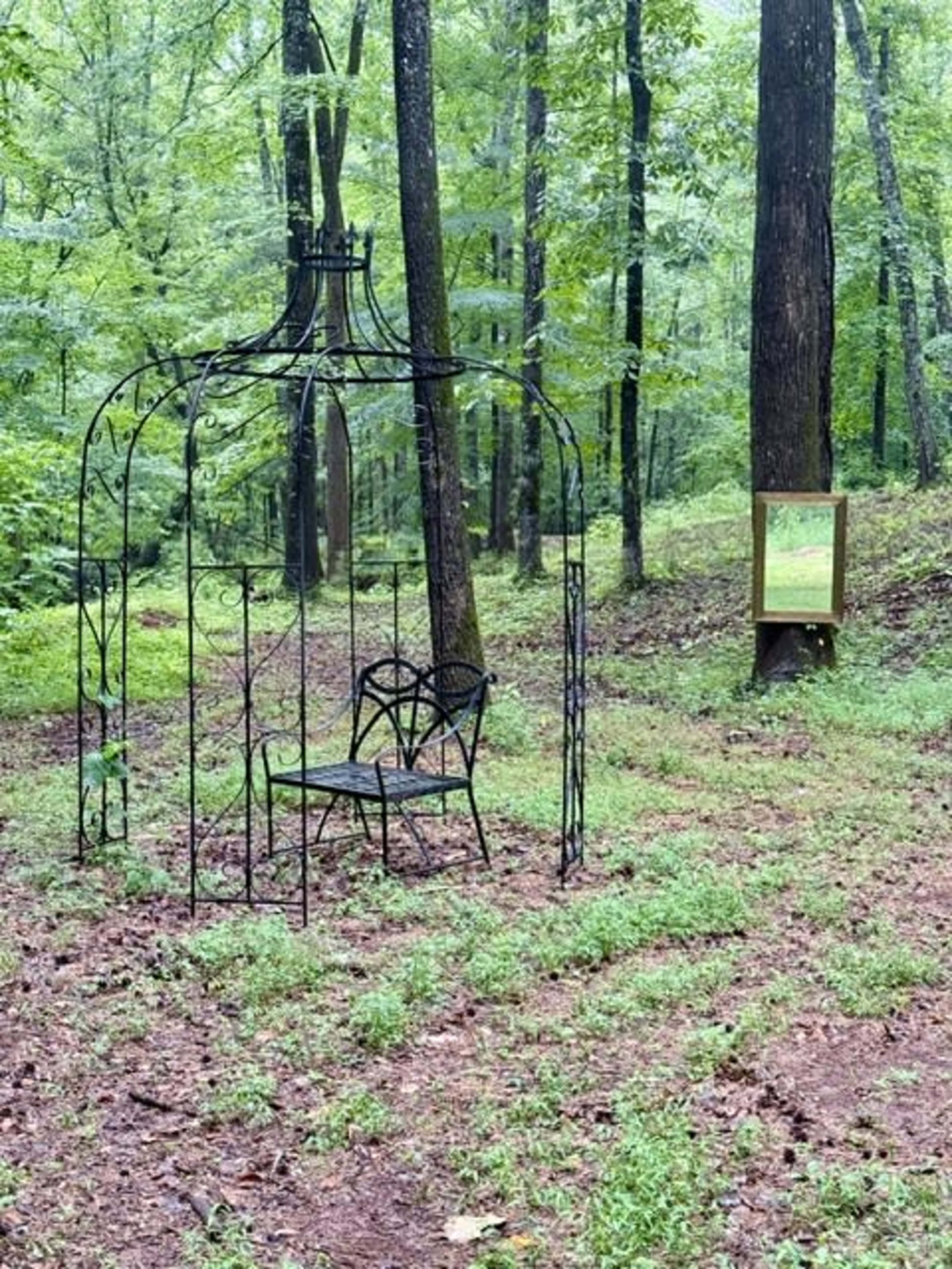 A wrought-iron structure with a bench is located in a wooded area with lush greenery and a framed sign nearby.
