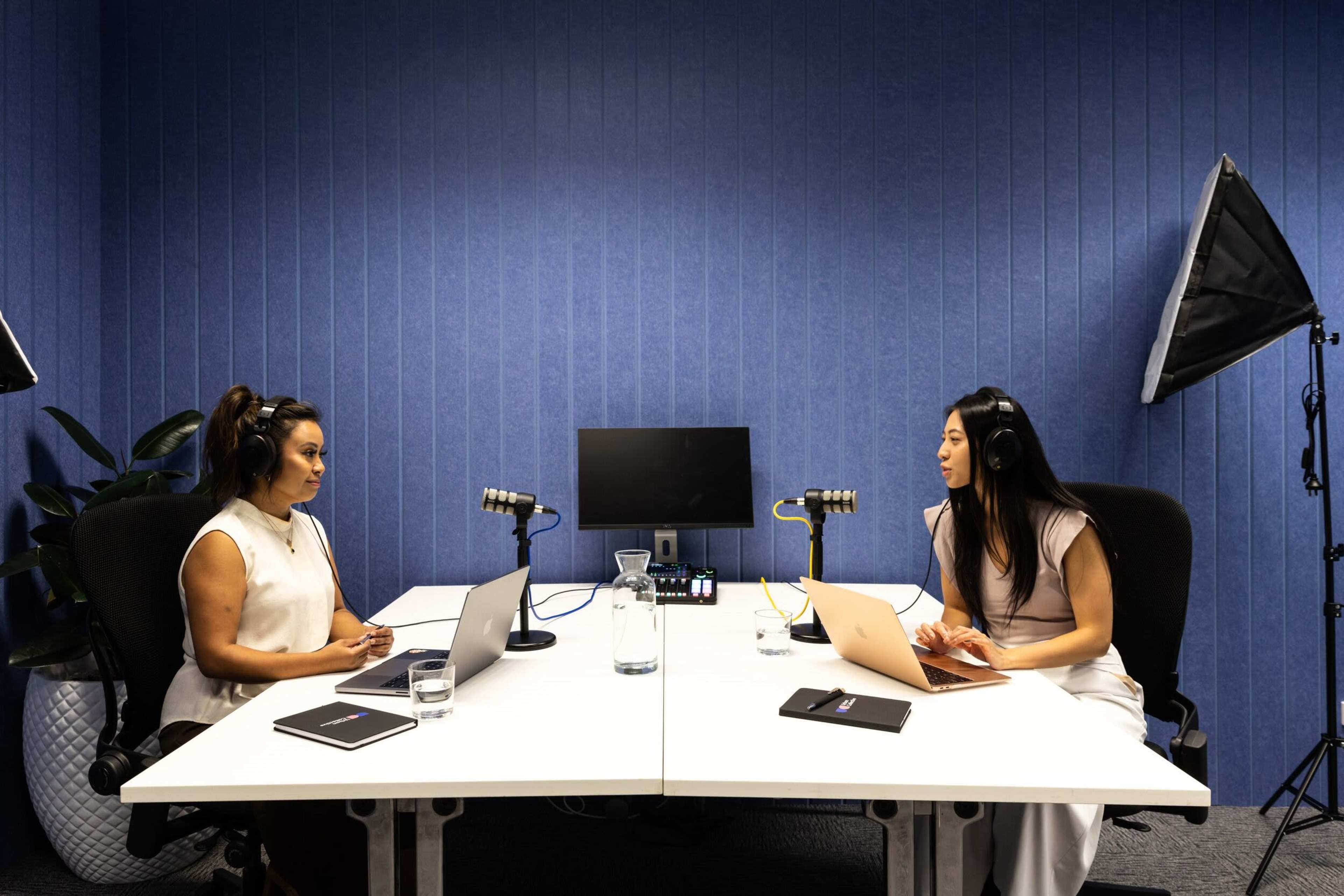 Two women are seated at a table in a blue-walled room, facing each other with laptops and microphones set up for a recording session.