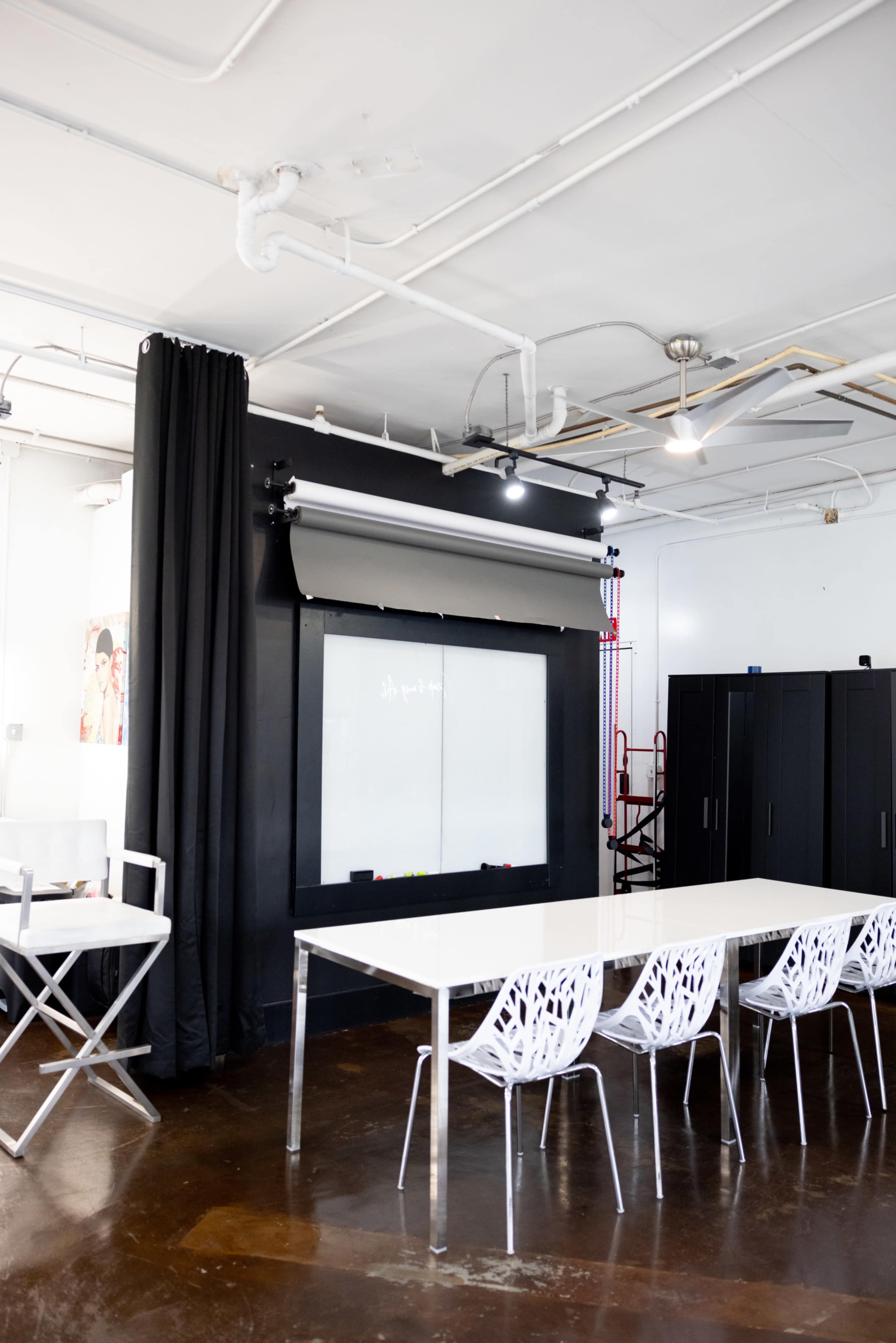 A modern conference room features a white table surrounded by transparent chairs, with a large projection screen on one wall and storage cabinets nearby.