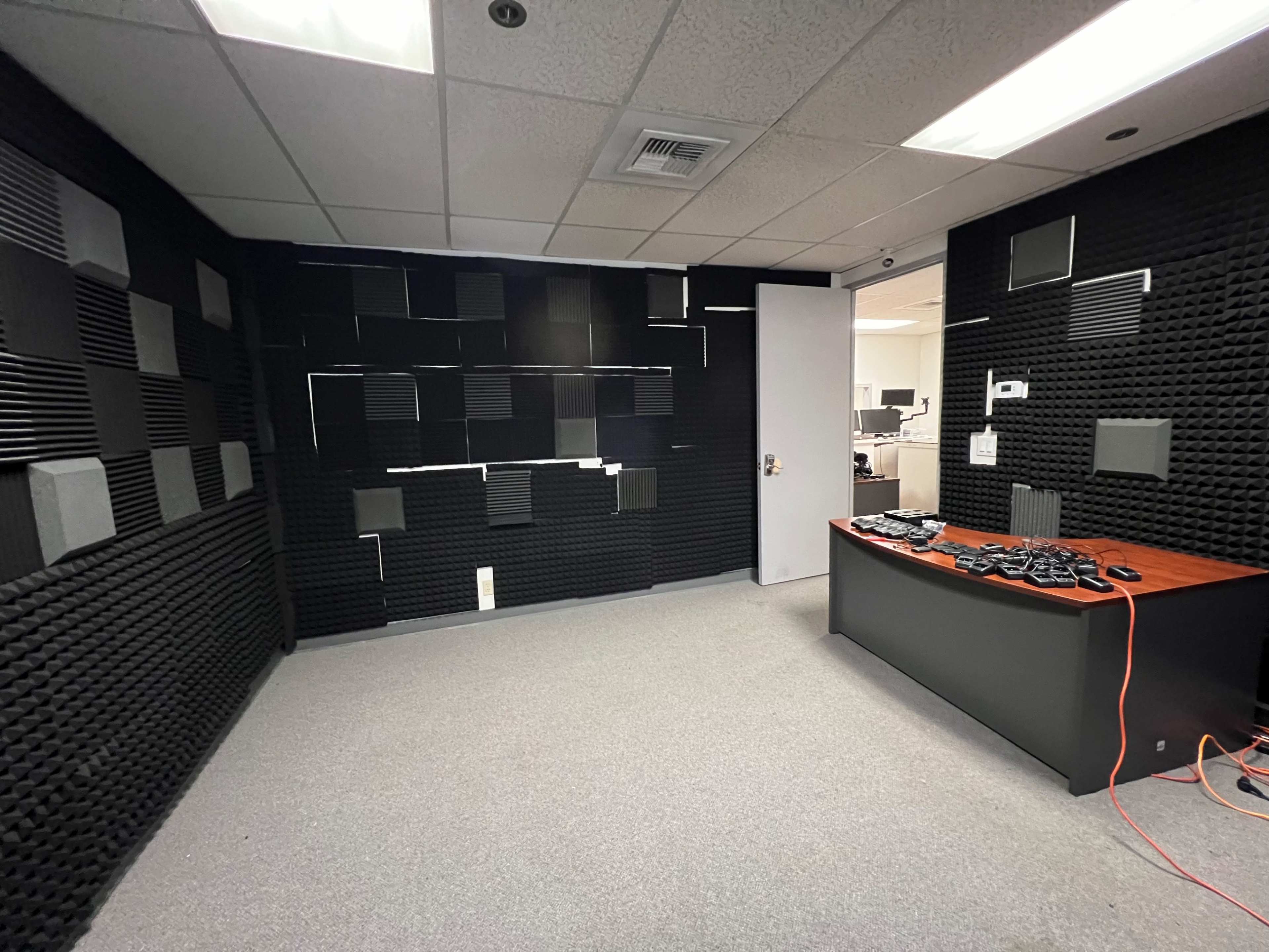 A soundproof room featuring black acoustic foam panels on the walls and a dark desk with electronic equipment on it.
