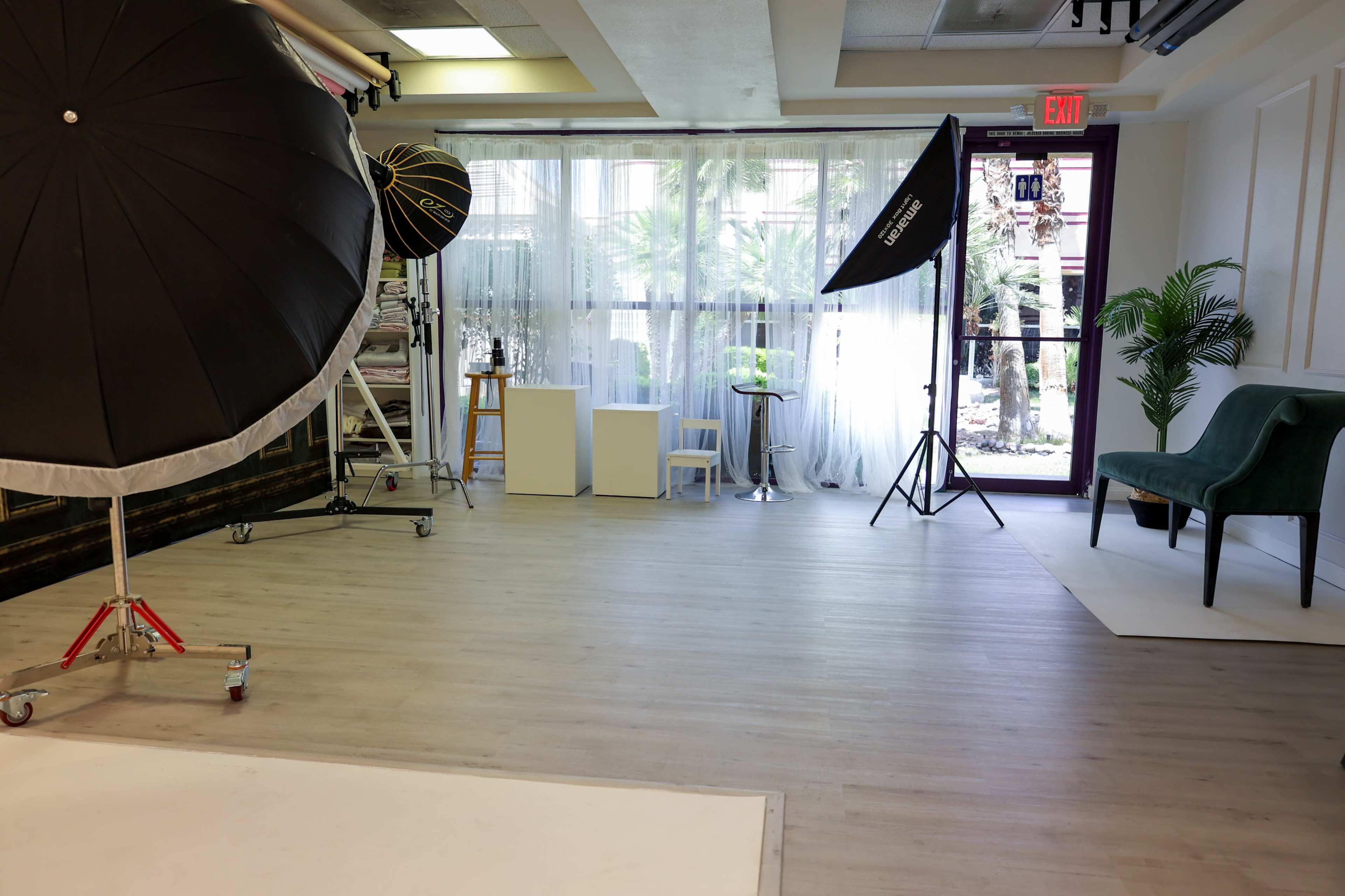 The image shows a bright and spacious photography studio equipped with various lighting equipment and seating areas, featuring large windows that allow natural light.