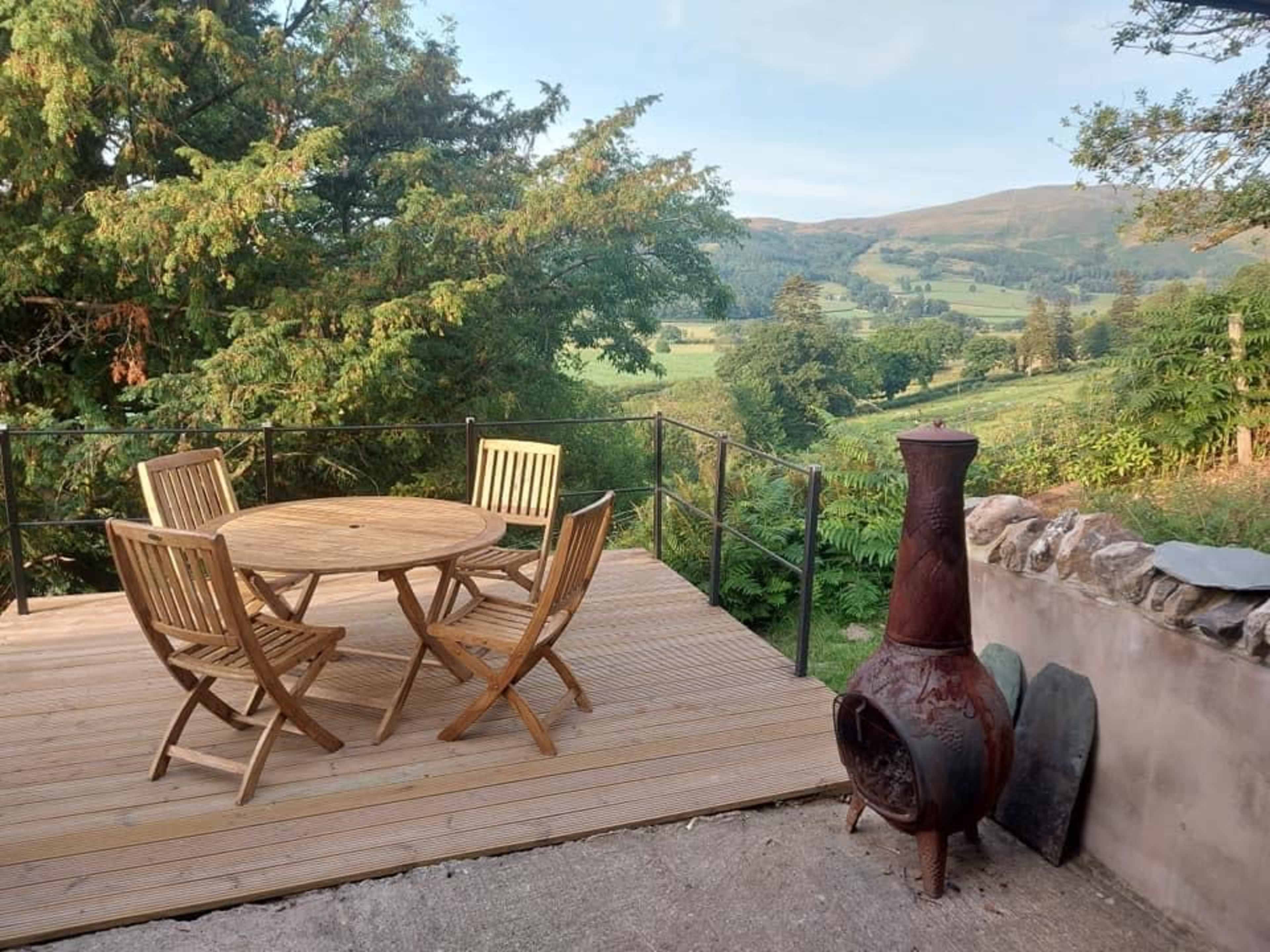A wooden deck with a round table and four chairs overlooks a green landscape and distant hills.
