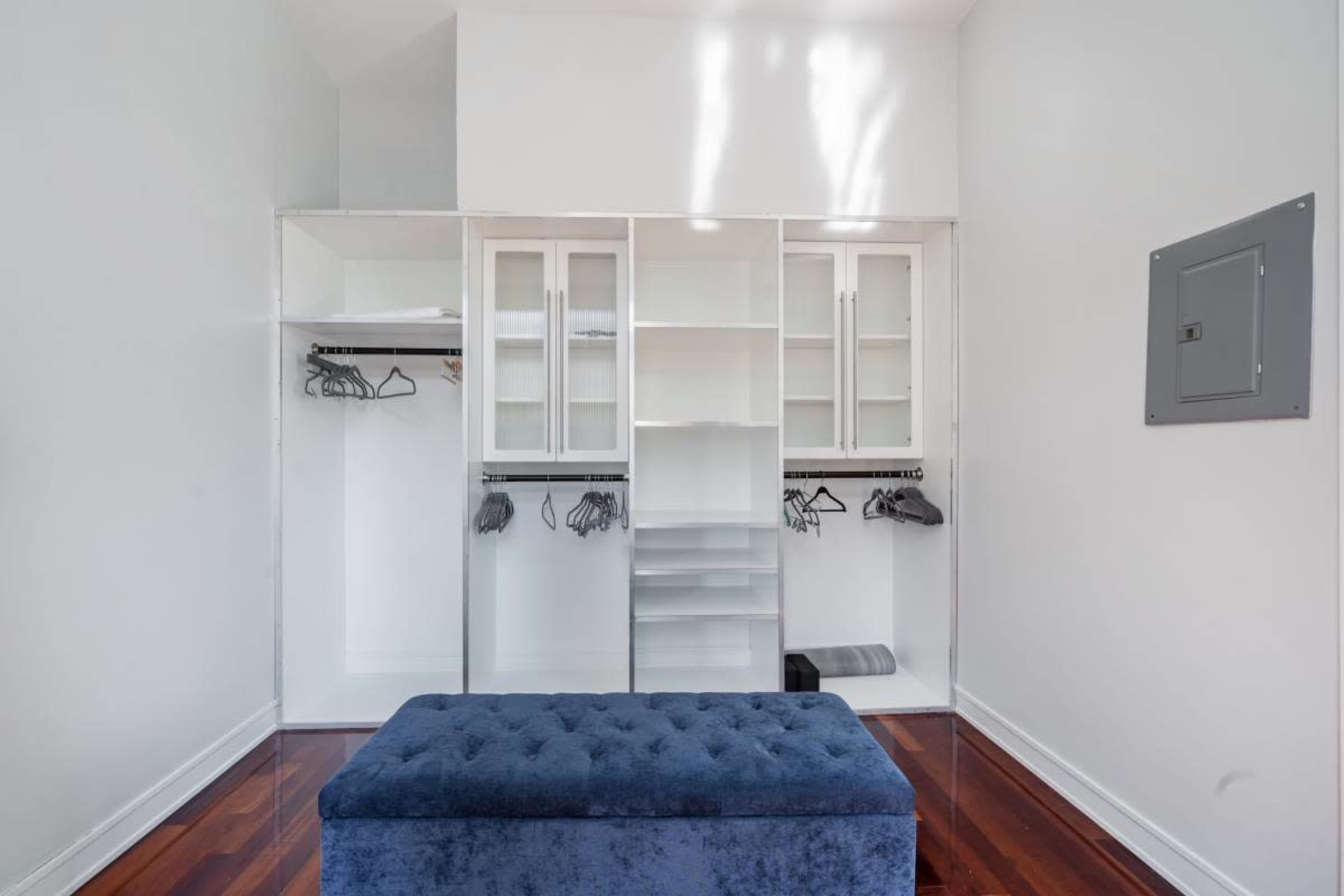 The image shows a spacious, empty closet with hanging racks, shelves, and a blue ottoman in front of it.