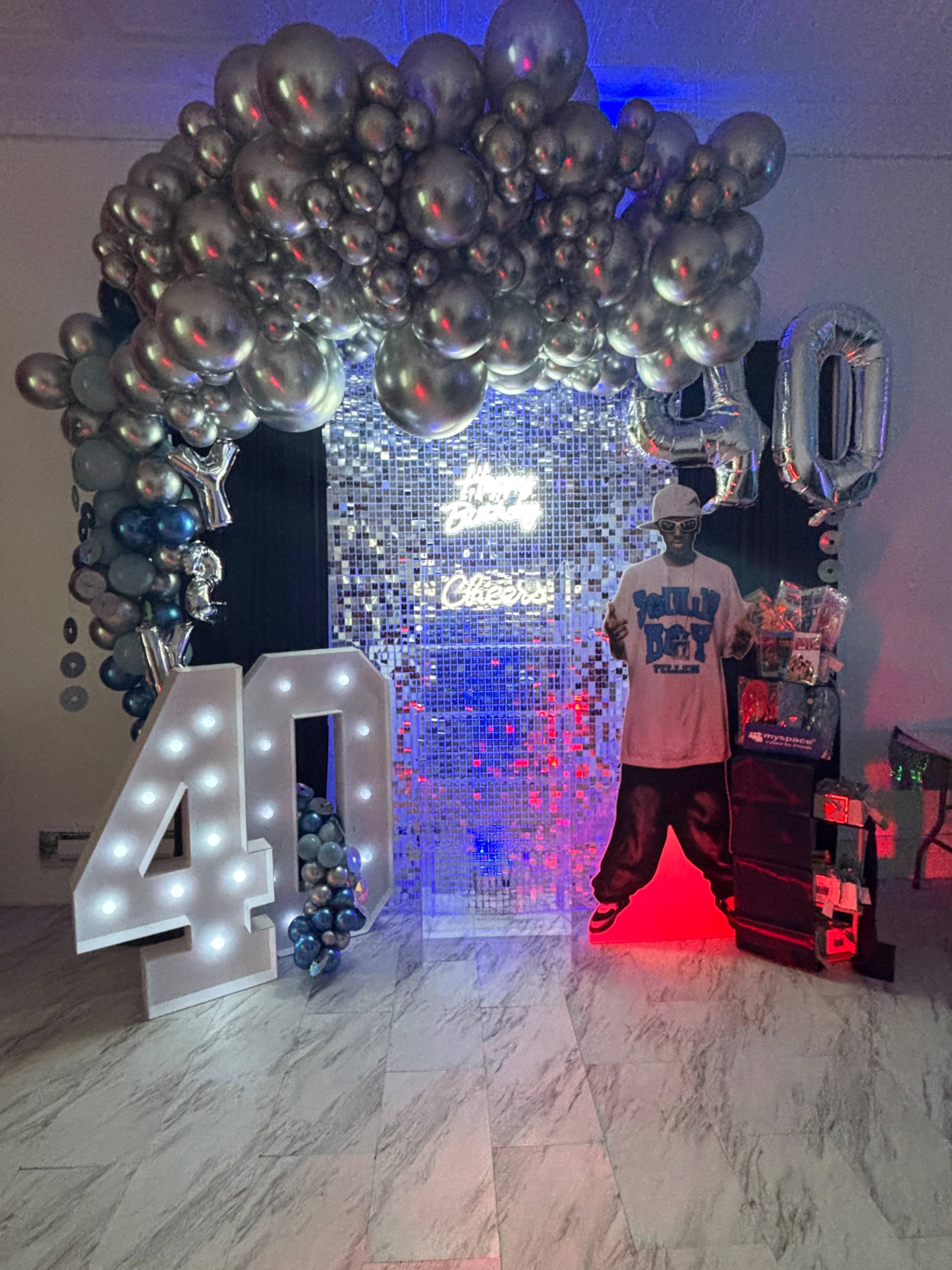 The image shows a decorative setup for a 40th birthday celebration, featuring a large "40" made of lights, a backdrop of mirrored tiles, balloons in silver and blue, and a figure dressed in a T-shirt with a party theme.
