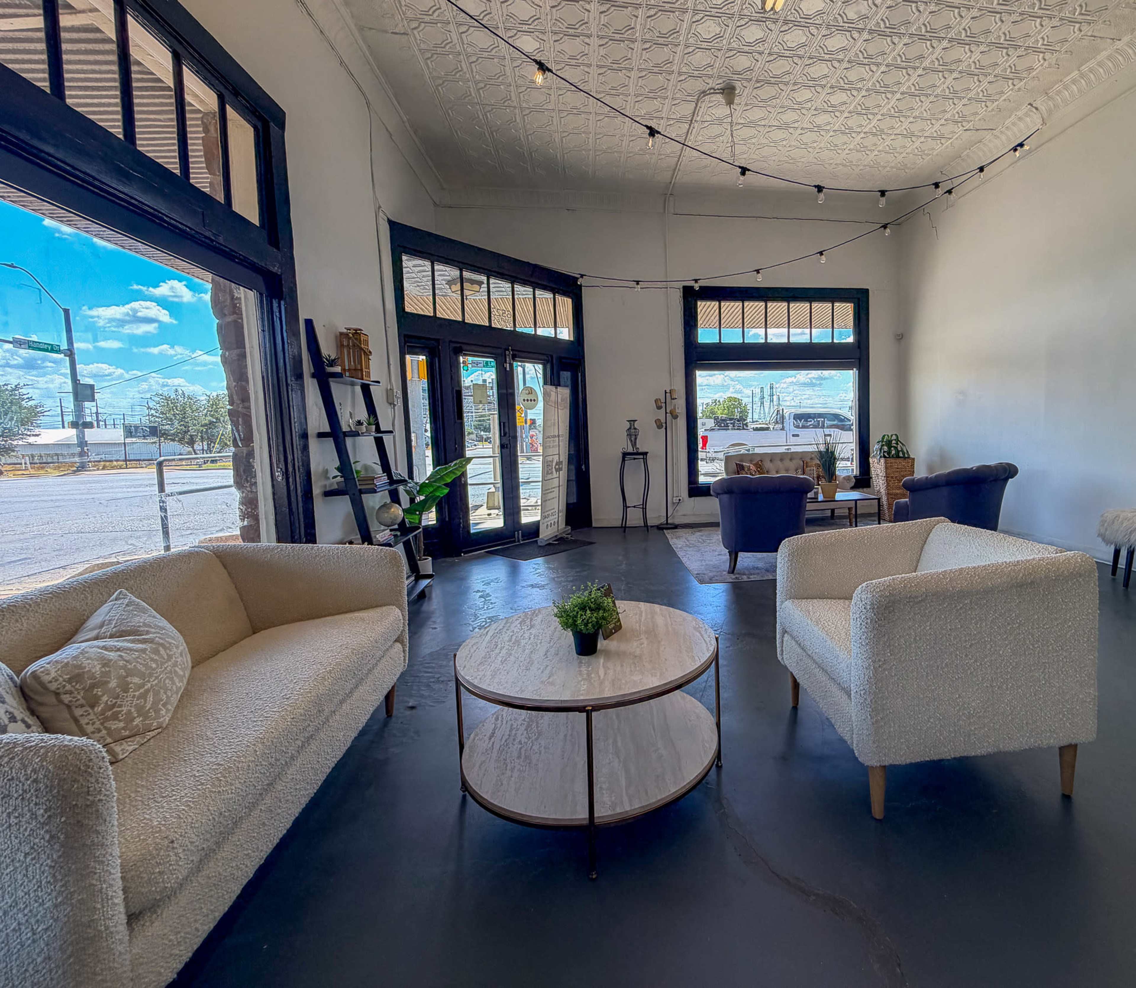 The image shows a bright, open space with two light-colored sofas, a round coffee table, and large windows displaying a view of the outside.