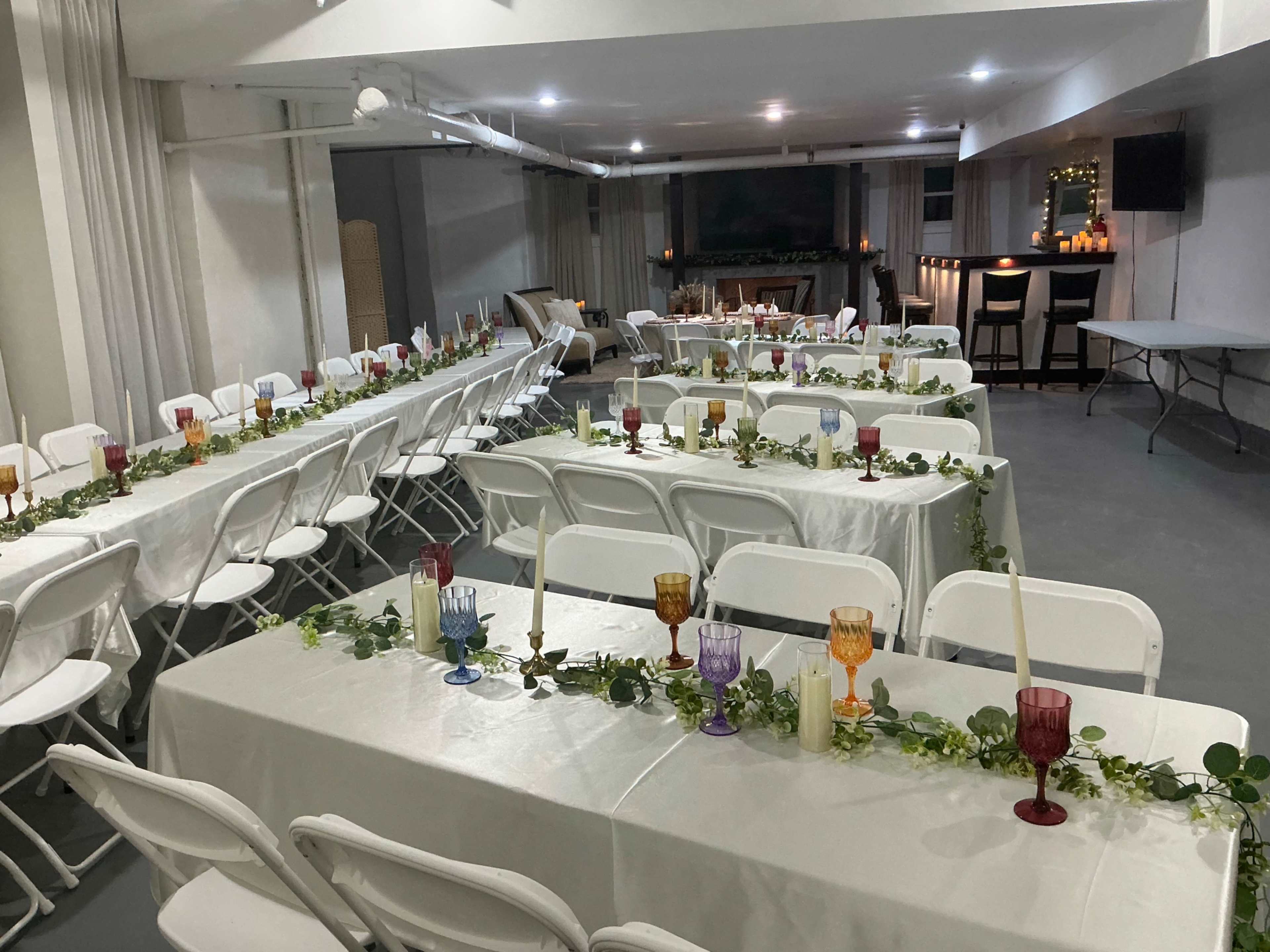 The image shows a moderately lit indoor venue arranged with rows of tables dressed in white tablecloths and decorated with greenery and colorful glasses.