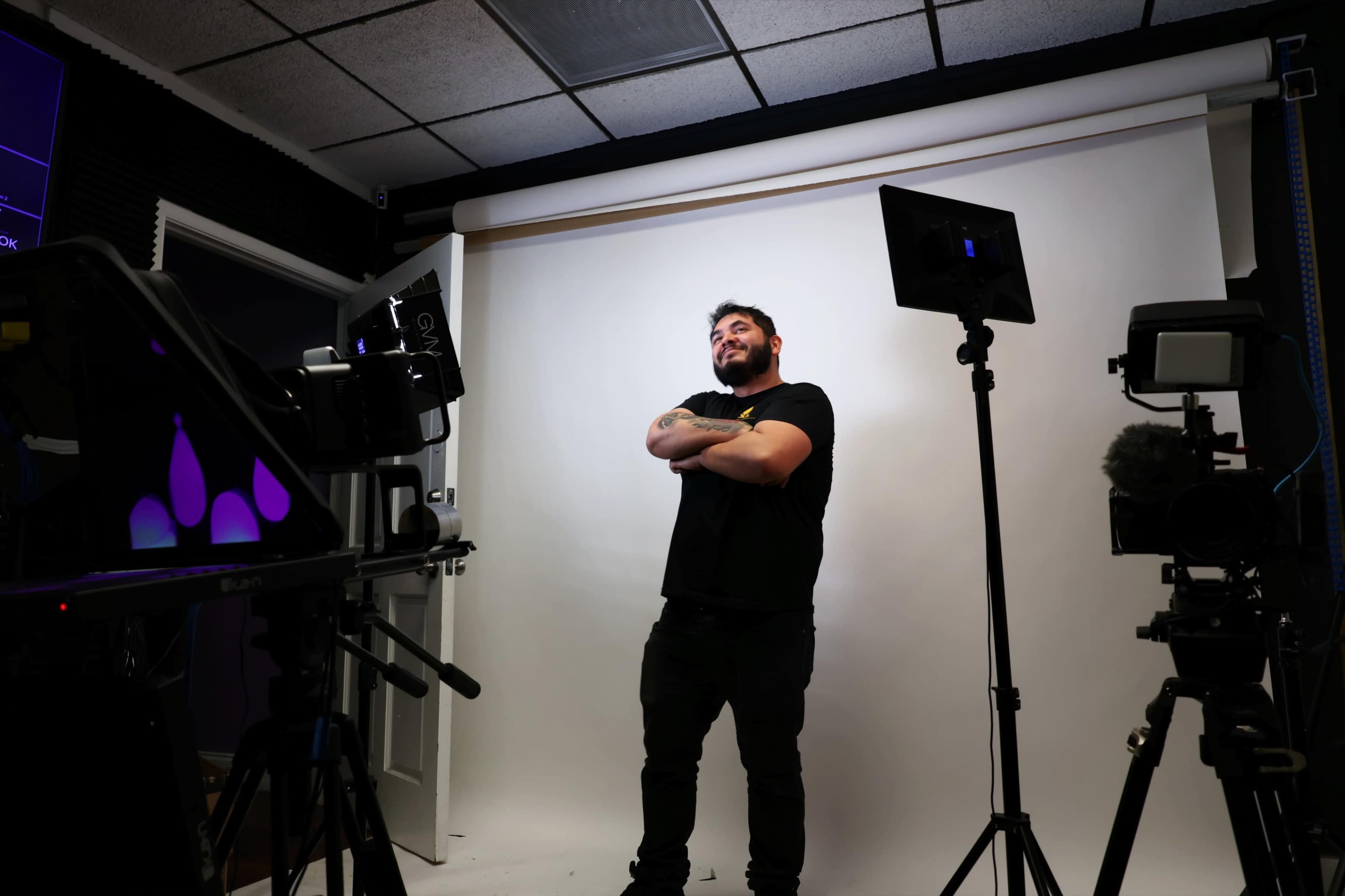 A man stands confidently in front of a plain white backdrop, flanked by video equipment and lights in a studio setting.