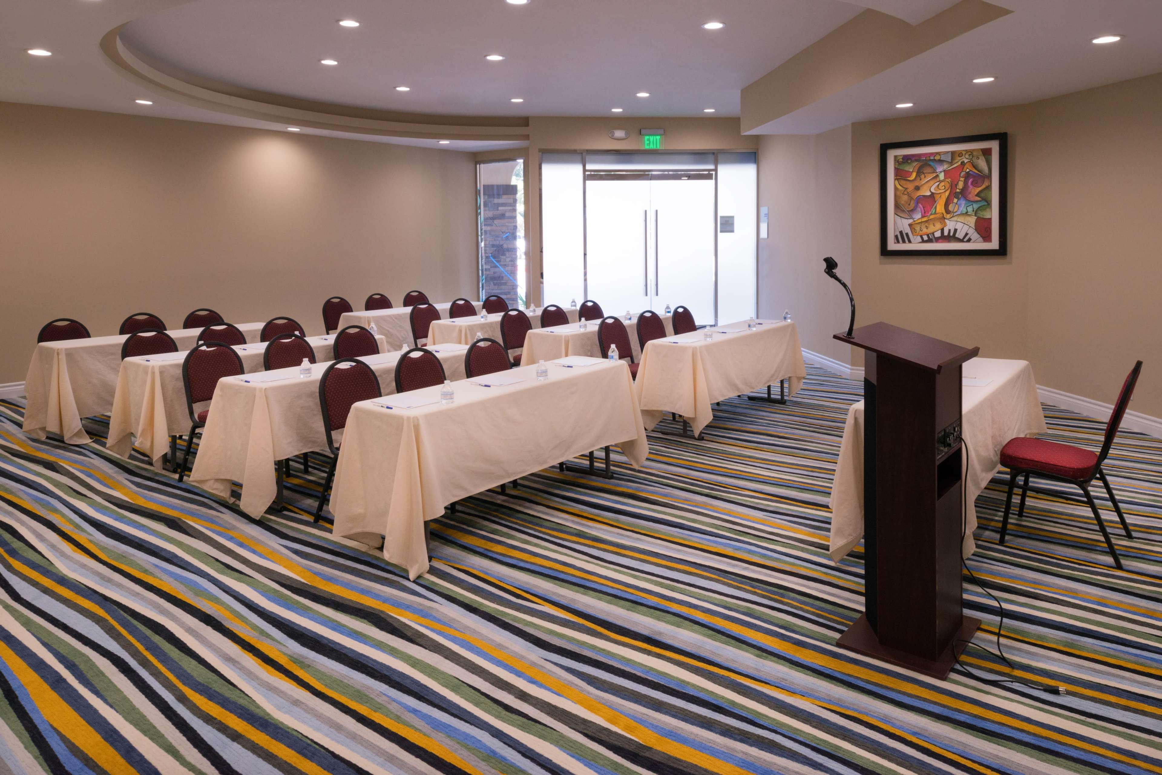 A conference room is set up with rows of tables and chairs, facing a podium against a backdrop of a colorful striped carpet.