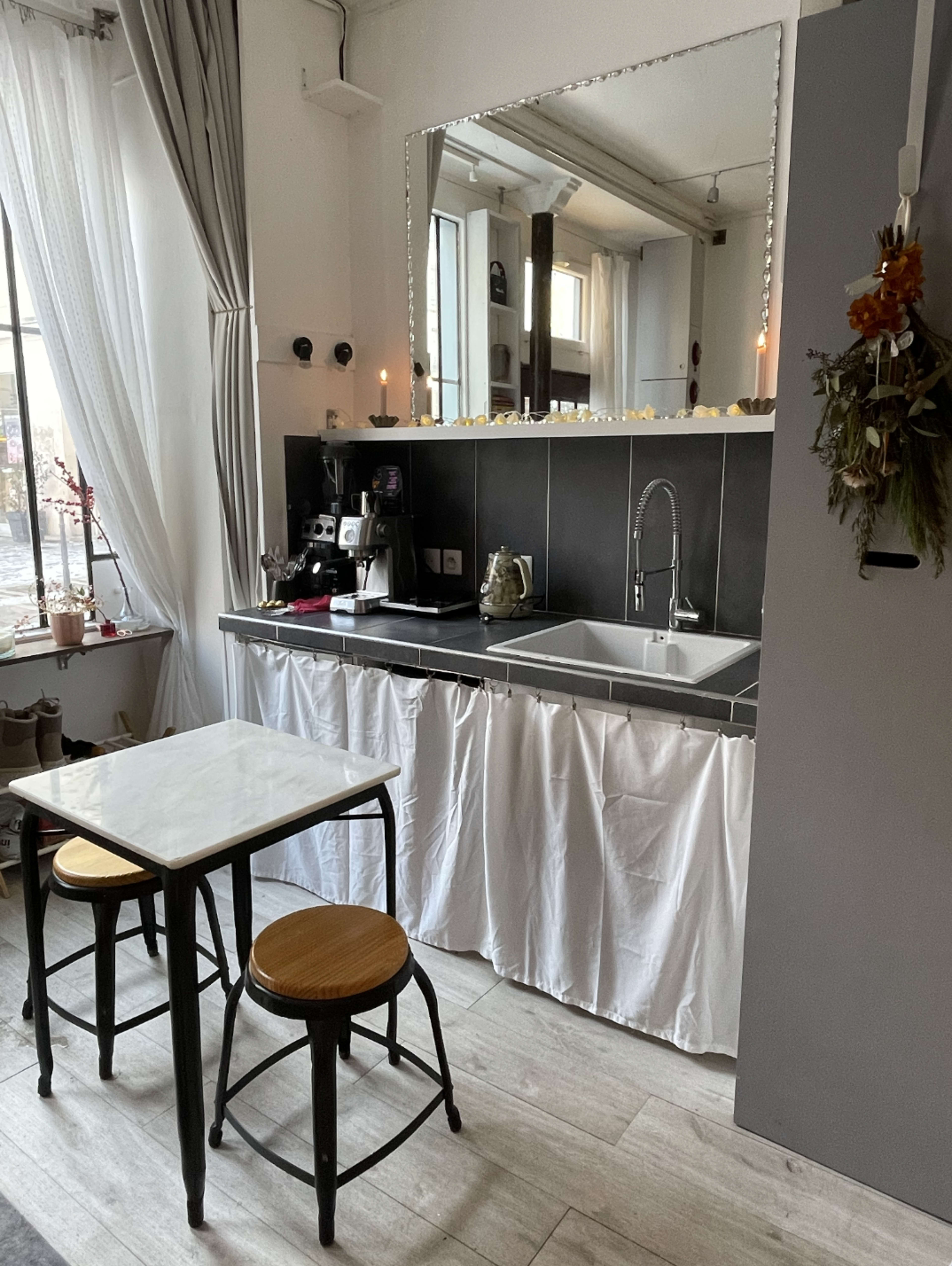 The image shows a compact kitchen area with a black countertop, a white curtain covering the space under the sink, two wooden stools, and a large mirror above the sink.