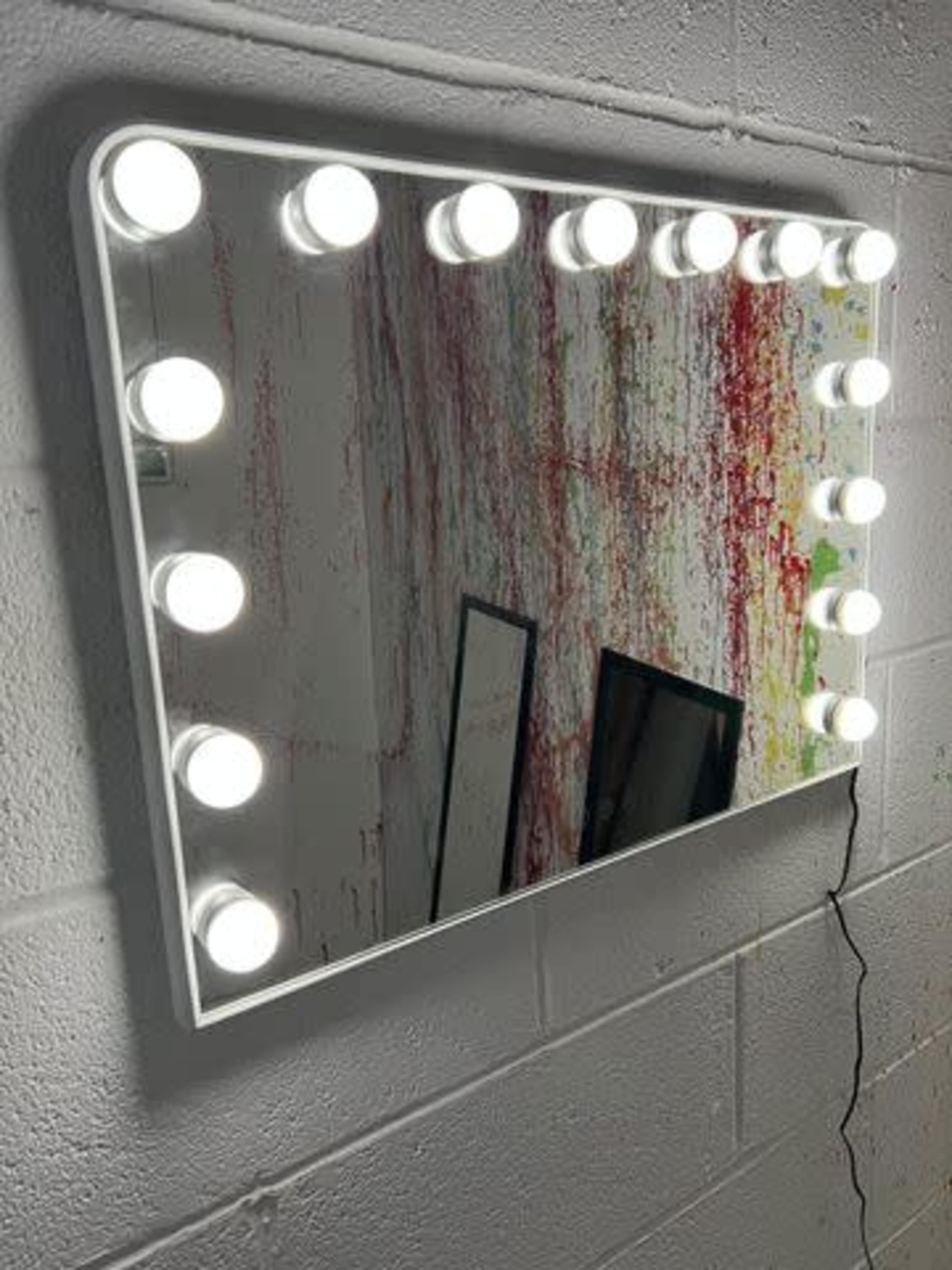 A rectangular mirror with bright circular lights is mounted on a wall featuring splashes of red paint.