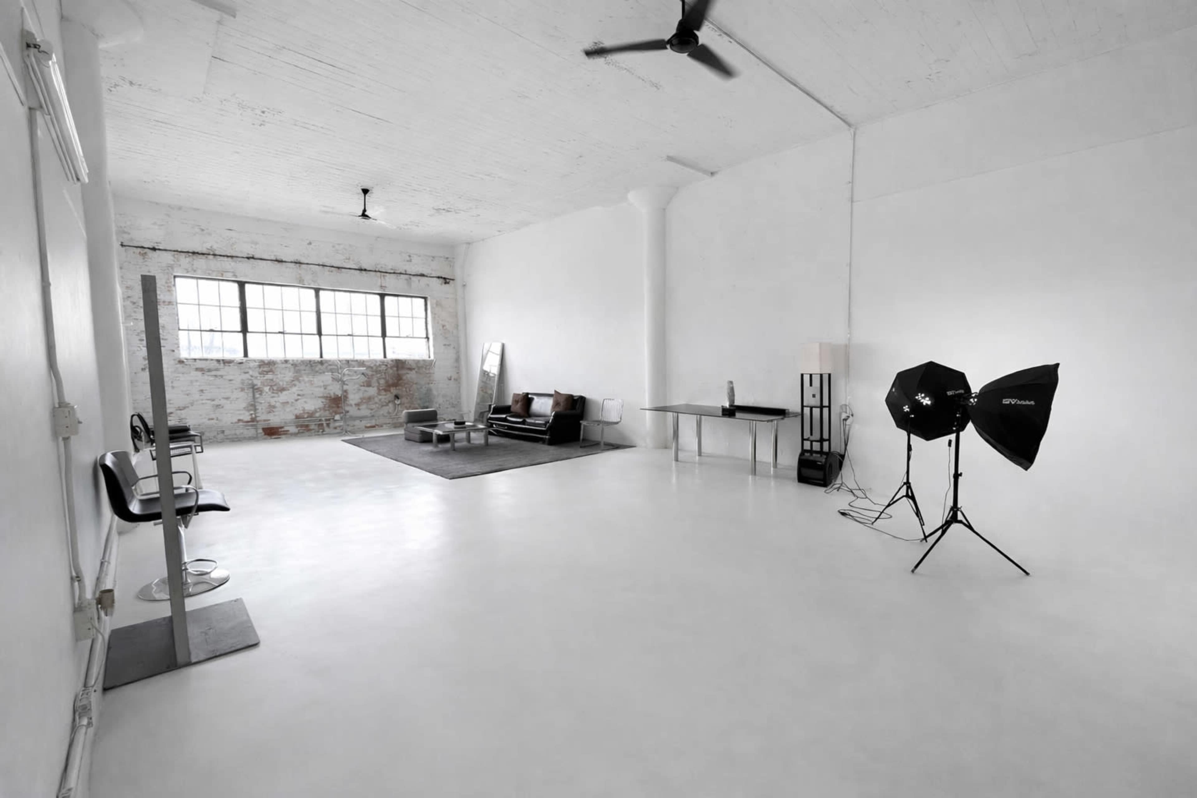 The image depicts a spacious, empty studio interior featuring white walls, a large window, minimalist furniture, and photography lighting equipment.
