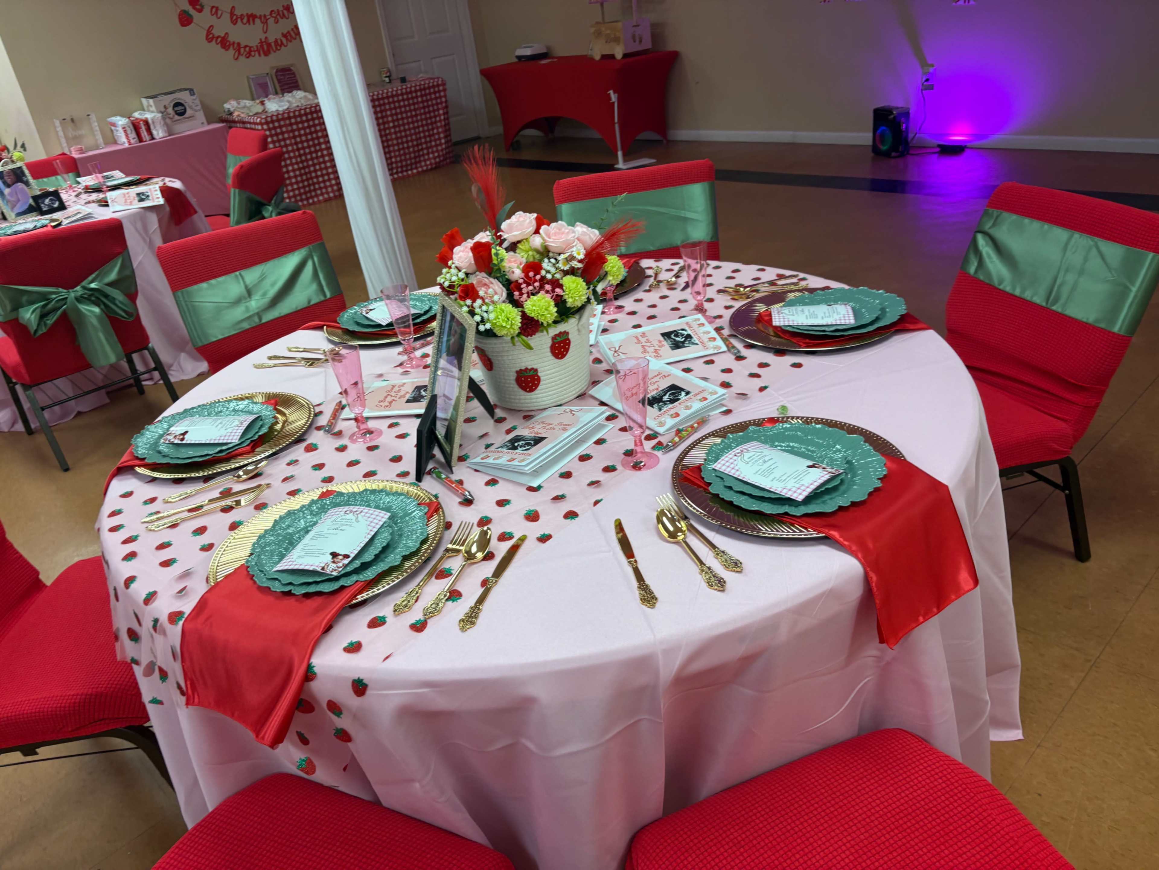 A round table is elegantly set with red and green decor, featuring floral centerpieces and vibrant tableware in a bright event space.