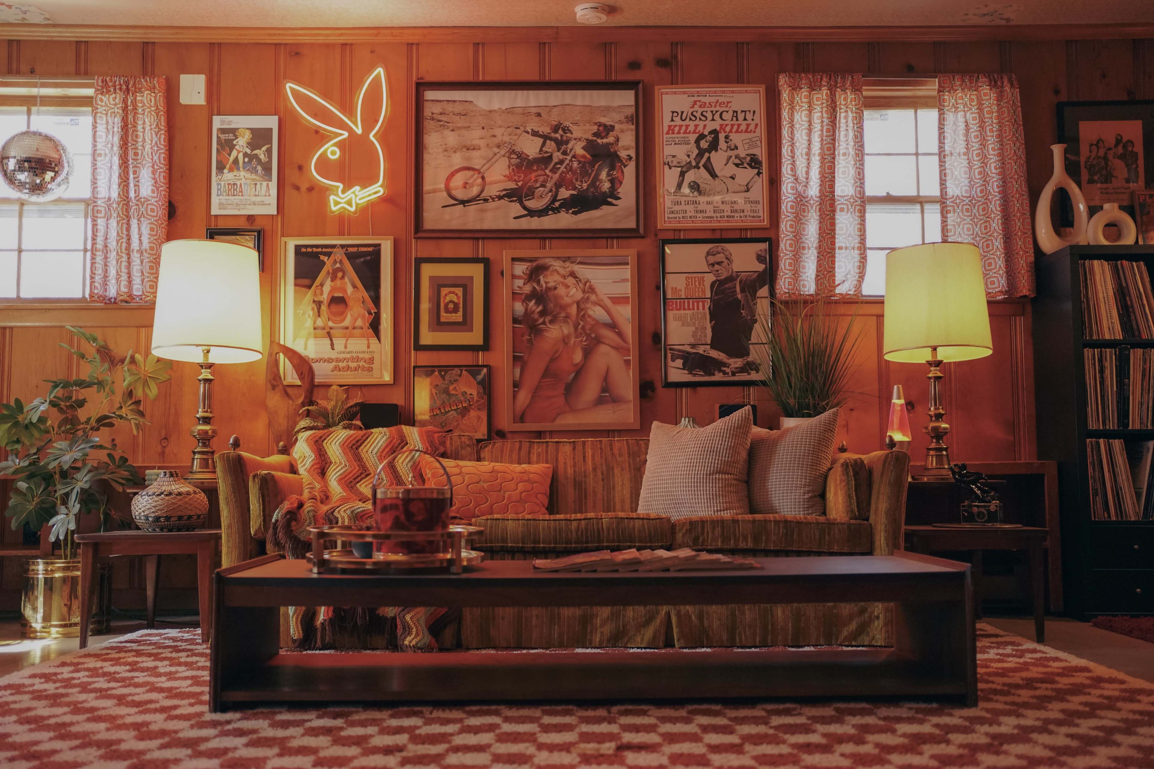 A cozy living room with wooden paneling, vintage posters on the walls, a neon Playboy sign, and a sofa with decorative cushions in front of a coffee table.