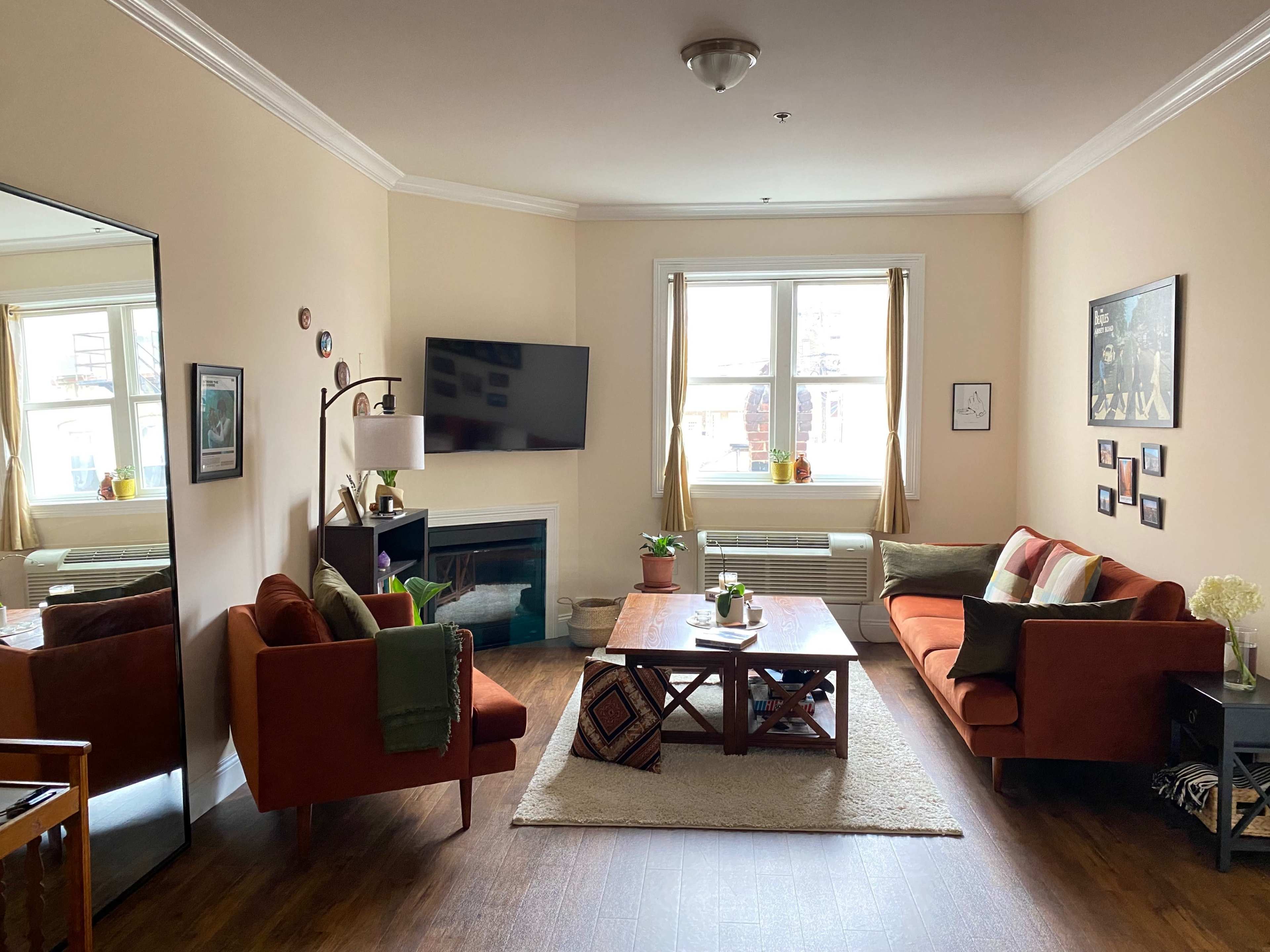 A cozy living room with two orange sofas, a wooden coffee table, and a fireplace beneath a television, all set against light-colored walls and large windows.