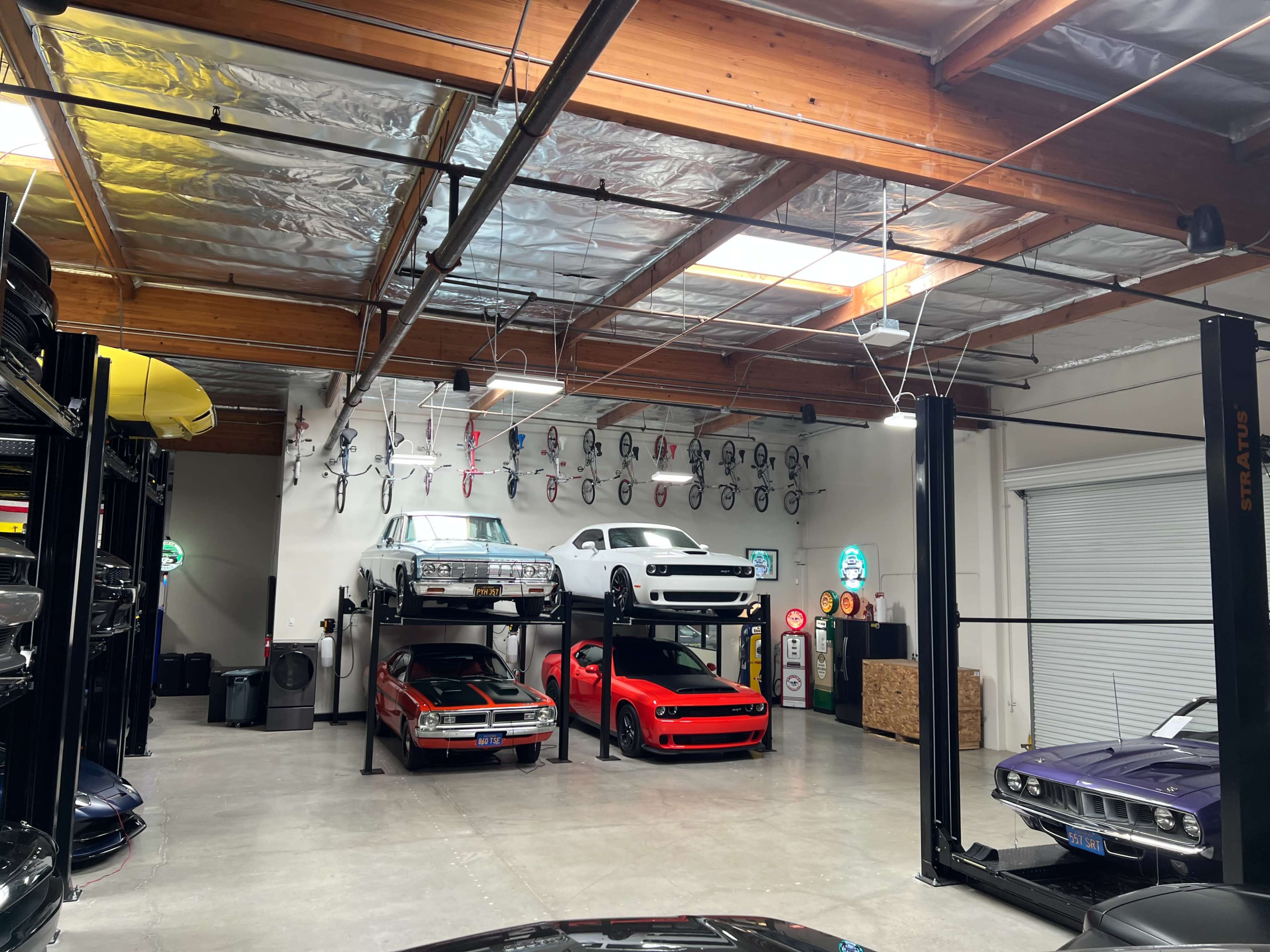 The image shows a spacious garage with two levels of car lifts, housing six cars including a classic muscle car and a sports car, along with bicycles mounted on the walls.