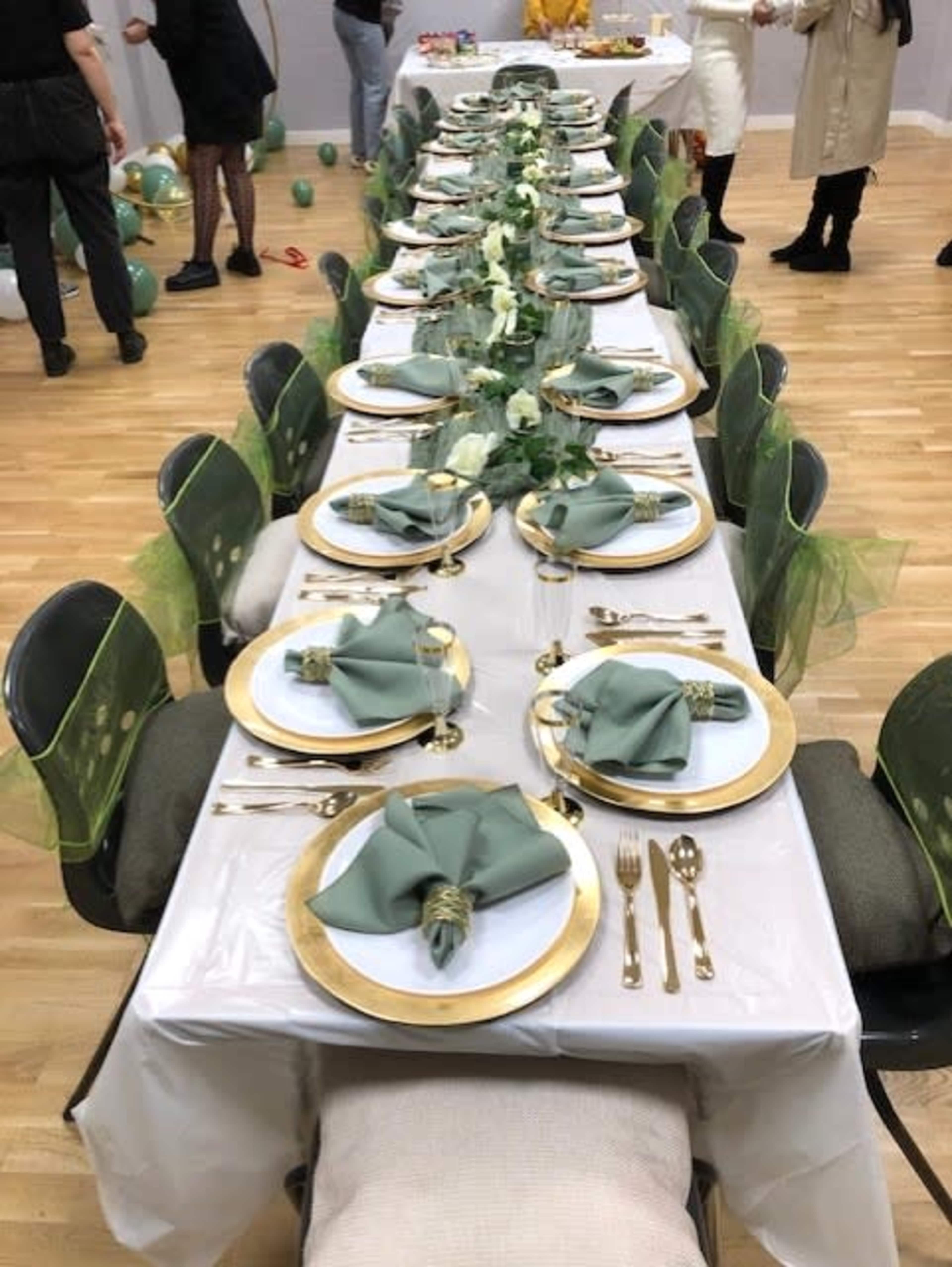 A long banquet table is set with green napkins, gold-rimmed plates, and golden cutlery, adorned with a floral centerpiece along the center.